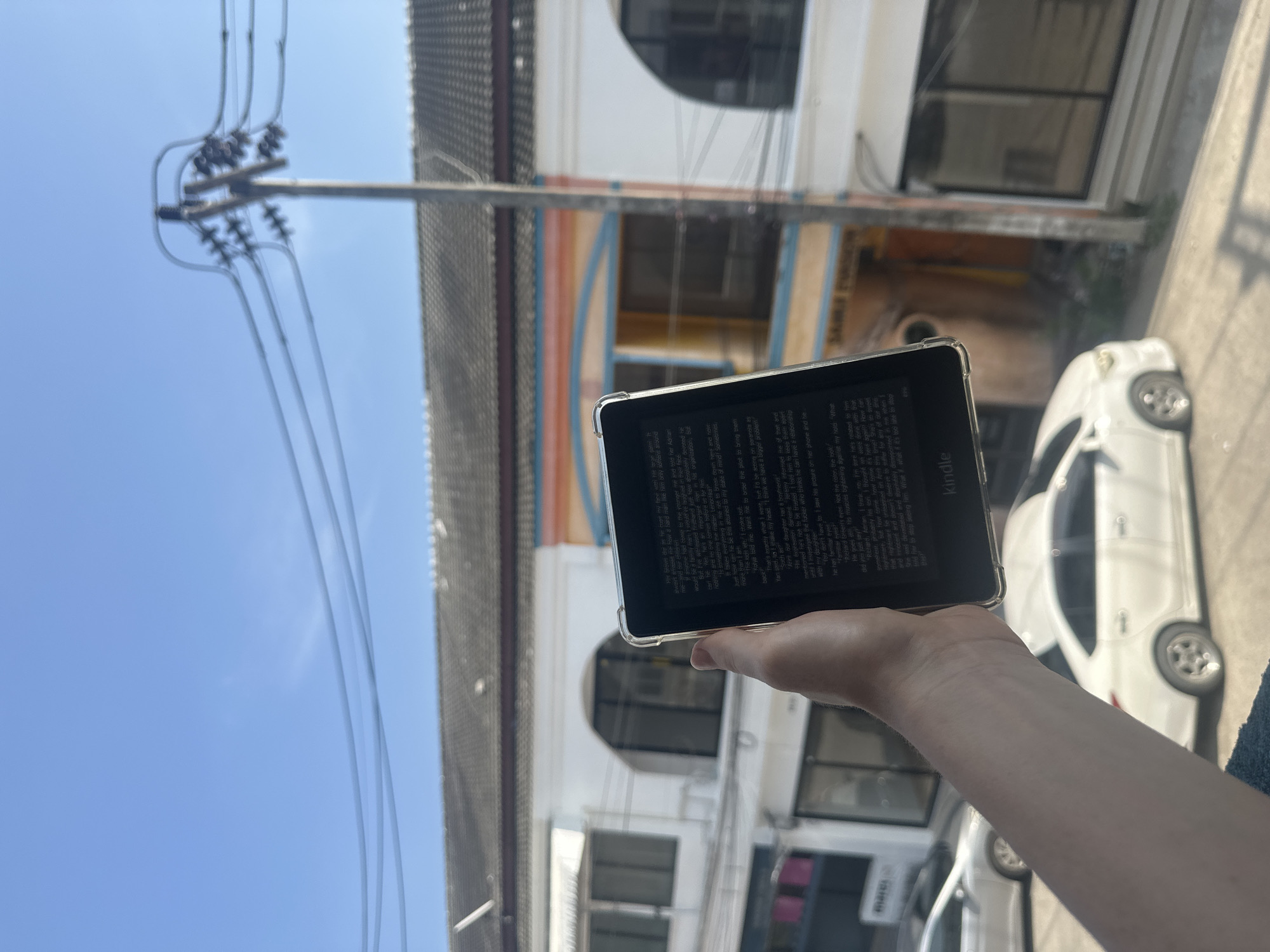 A person holds a black Kindle e-reader outdoors. The Kindle displays text. The background features a cityscape, including buildings with a white facade and arched windows. Power lines are visible against a blue sky. A white car is parked on the side of the road. The setting appears to be Baan Chaweng, Thailand.