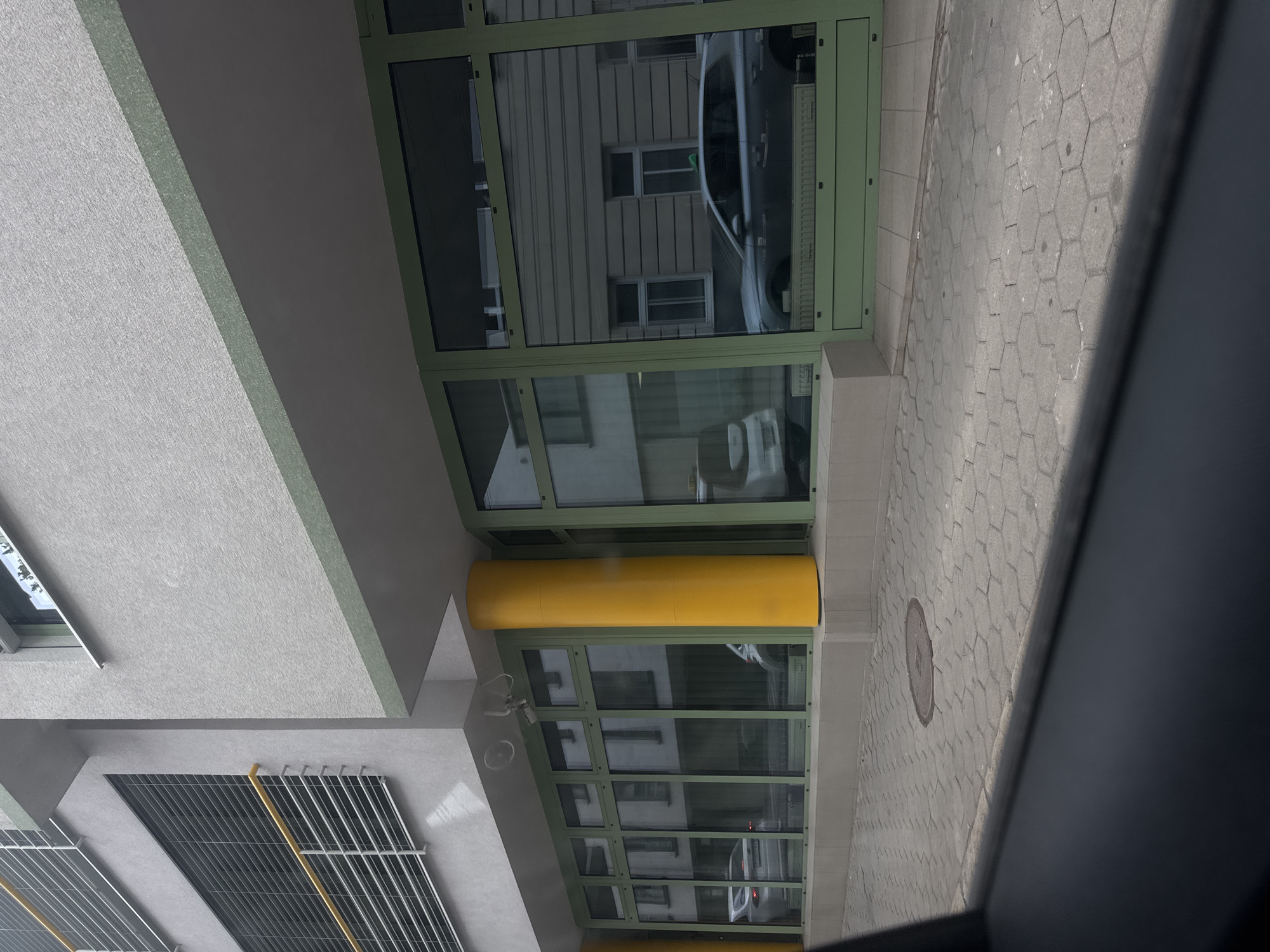 The image captures a modern building facade in Bratislava, Slovakia. The building features large, green-framed windows reflecting an adjacent building. A bright yellow cylindrical pillar is prominently visible, supporting an overhang. The ground in front of the building is paved with hexagonal interlocking stones. Two vehicles are visible through the windows; one appears to be a silver sedan and the other is a white car. A circular manhole cover is present on the pavement. The overall scene suggests an urban commercial or office building setting.