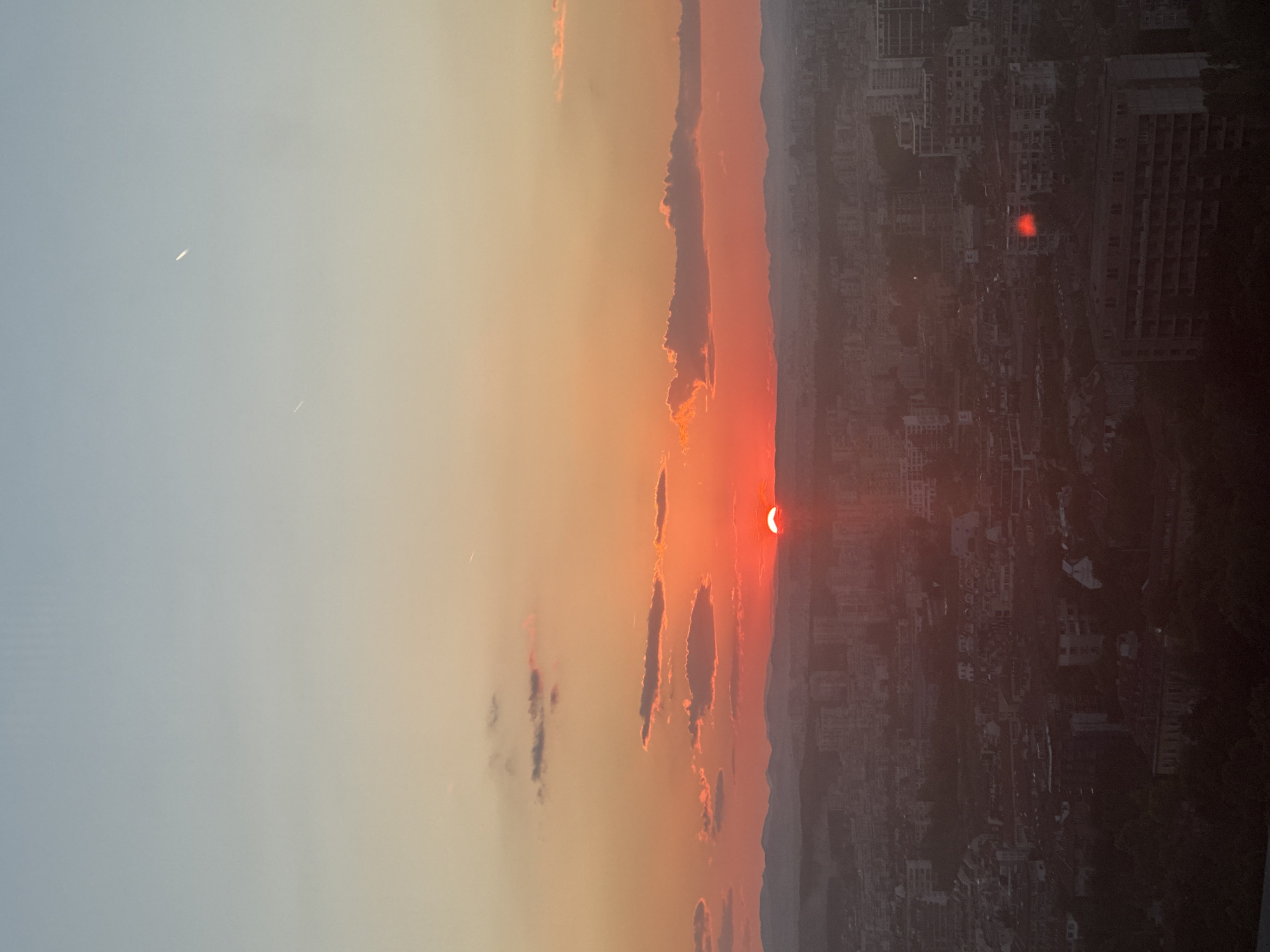 This is a photograph of a sunset over the city of Sofia, Bulgaria. The sun is a bright red orb, partially obscured by a hazy horizon. The sky transitions from a pale blue at the top to a vibrant orange and red near the horizon, with scattered clouds catching the last rays of sunlight. In the distance, a mountain range is visible. The foreground is dominated by a dense urban landscape, with numerous buildings and structures stretching out to the horizon. Some of the buildings appear to be residential, while others seem to be commercial or industrial. The overall impression is of a city settling down for the evening, bathed in the warm glow of the setting sun. There are no people or specific actions depicted in the image, it is a static landscape view.