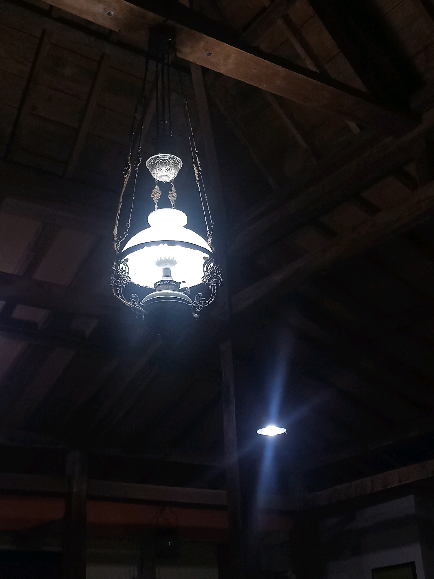 An antique kerosene lamp hangs from the wooden ceiling of a building in Bakalanpule, Indonesia. The lamp has a white glass shade and intricate metalwork. A second, smaller light fixture is visible in the background, casting a blueish glow. The room appears dimly lit, with exposed wooden beams supporting the ceiling. No people are present in the scene.