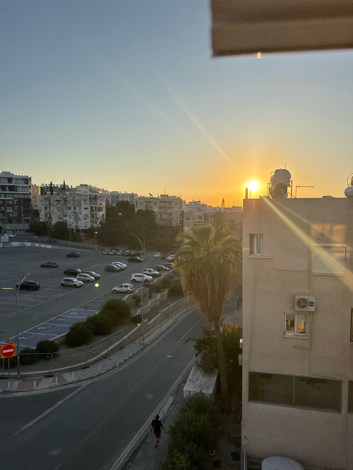 The image depicts an urban scene at sunset in Agios Tychonas Community, Cyprus. The sun is low on the horizon, casting a warm orange glow across the sky and creating lens flares that streak across the frame. In the foreground, a light-colored building with several windows and an air conditioning unit is visible on the right. A palm tree stands to the left of this building. Beyond the buildings, a large, mostly empty parking lot is visible, with several cars parked in designated spots. A road runs alongside the parking lot, and a lone pedestrian is walking on the sidewalk. In the background, a cluster of apartment buildings and other structures stretches across the landscape, bathed in the soft evening light. The overall impression is one of a quiet urban environment at the end of the day.