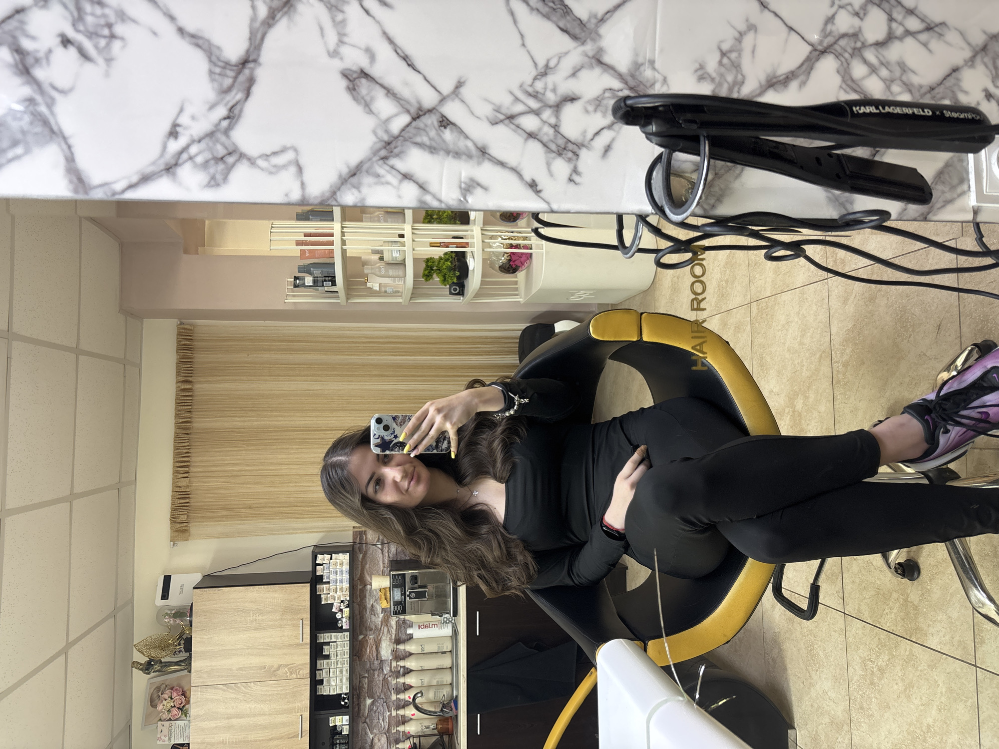 A young woman is captured in a mirror selfie, seated in a salon chair within what appears to be a hair salon in Chirpan, Bulgaria. She has long, wavy brown hair, wears a black long-sleeved top, black leggings, and purple and black athletic shoes. Her left hand, adorned with yellow nail polish, holds a smartphone with a patterned case, angled to take the picture. She smiles softly at her reflection.
The setting is an indoor space with a tiled floor and neutral-toned walls. Reflected in the mirror, behind the woman, is a white marble-patterned surface. On this surface rests a black hair straightener clearly labeled "KARL LAGERFELD x SteamPod". Below the marble, various shelves display numerous hair products, including shampoo and conditioner bottles, and small decorative items like green plants and pink flowers in vases. A partial view of a black display unit holding rows of what appear to be hair dye samples or swatches is visible. Another section shows wooden cabinets. The floor in front of the chair has text partially visible, spelling out "HAIR ROOM". A beige textured curtain or blind can also be seen in the background reflection. The salon appears well-lit, suggesting daytime or bright artificial lighting.