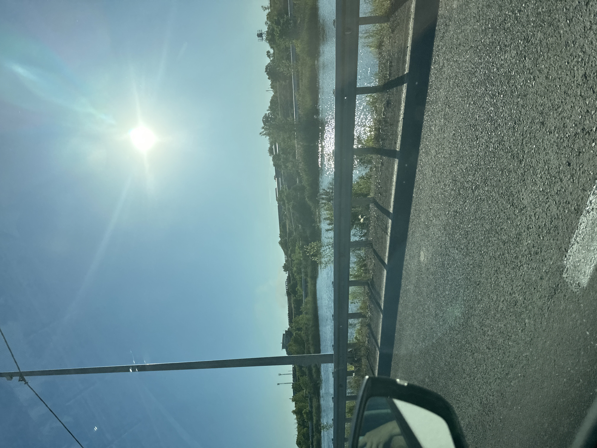 This is a vertical, eye-level, outdoor shot taken from a vehicle, possibly in Abakan, Russia. The scene depicts a roadside view on a sunny day. The sun is prominently featured in the upper left quadrant, casting a bright glare and lens flare across the clear blue sky. To the right of the sun, a metal guardrail runs horizontally, separating the road from a body of water. The water, reflecting the sunlight, appears calm and is bordered by lush green vegetation and trees that extend up a gentle incline. In the distance, on the far side of the water, there are some structures visible, including what appears to be a water tower or industrial building. The road itself is visible in the foreground, with its asphalt texture clearly rendered, and a portion of a car's side mirror is in the lower right corner. A tall metal pole, possibly a lamppost, stands vertically on the left side of the frame. The overall impression is a peaceful, bright daytime scene, likely during the warmer months given the foliage. There is no visible text in the image.