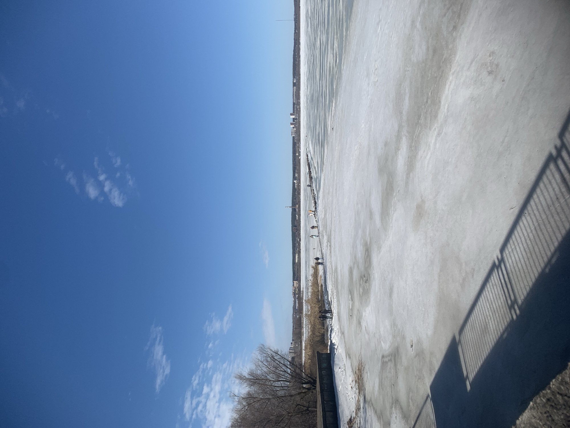 The image shows a frozen pond in Izhevsk, Russia. The sky is blue with a few scattered clouds. In the distance, there is a city skyline with a tall tower. The pond is covered with ice and a few people are walking on it. The shadow of a fence can be seen.