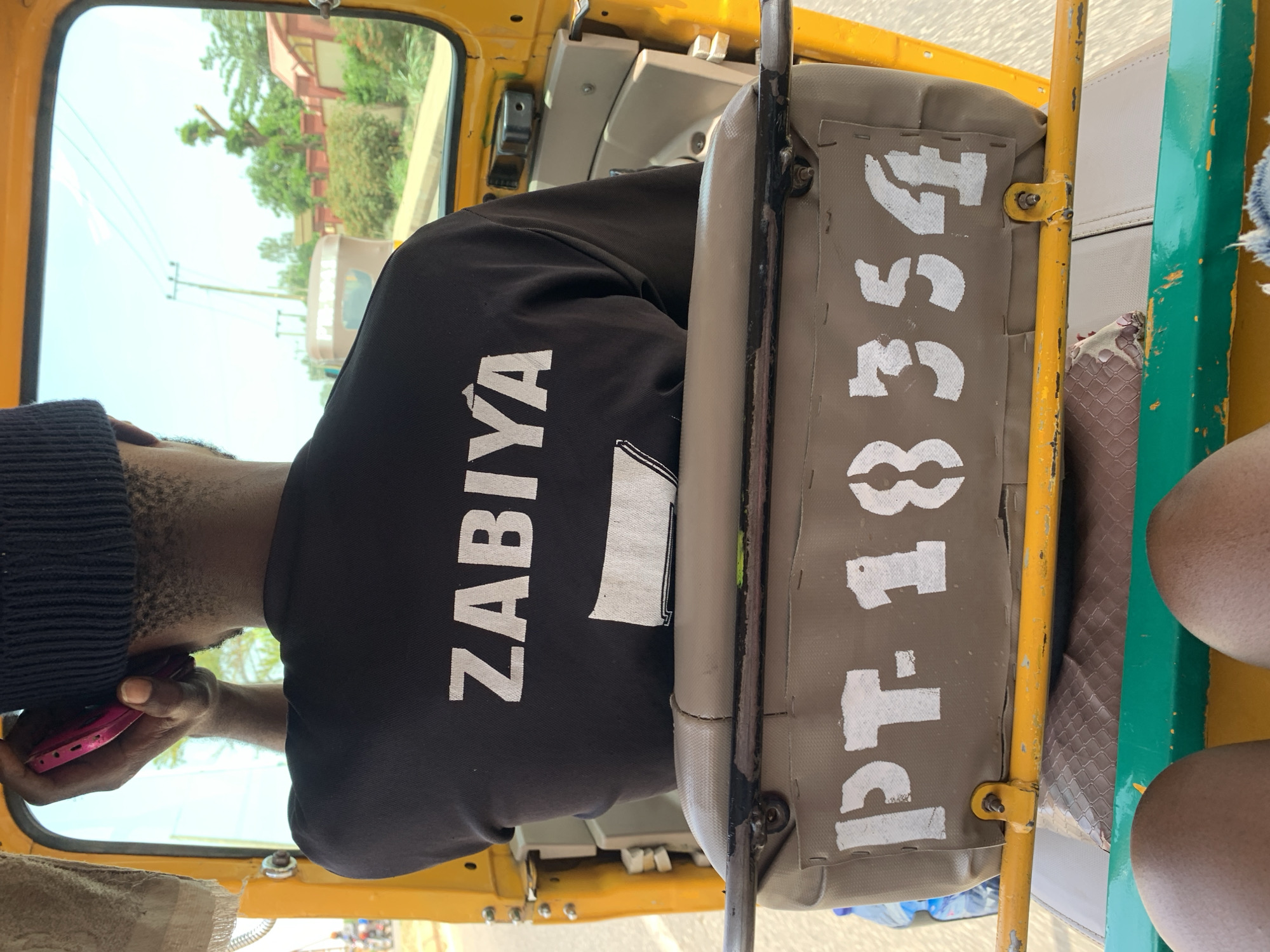 View from inside a commercial auto rickshaw in Rafin Par, Nigeria. A passenger wearing a black shirt with the name "ZABIYA" printed in white and a black knit cap is looking at a pink mobile phone. The vehicle seat in front of the passenger has the text "PT-18354" stenciled on the back in white letters. In the background, trees and buildings are visible through the window.