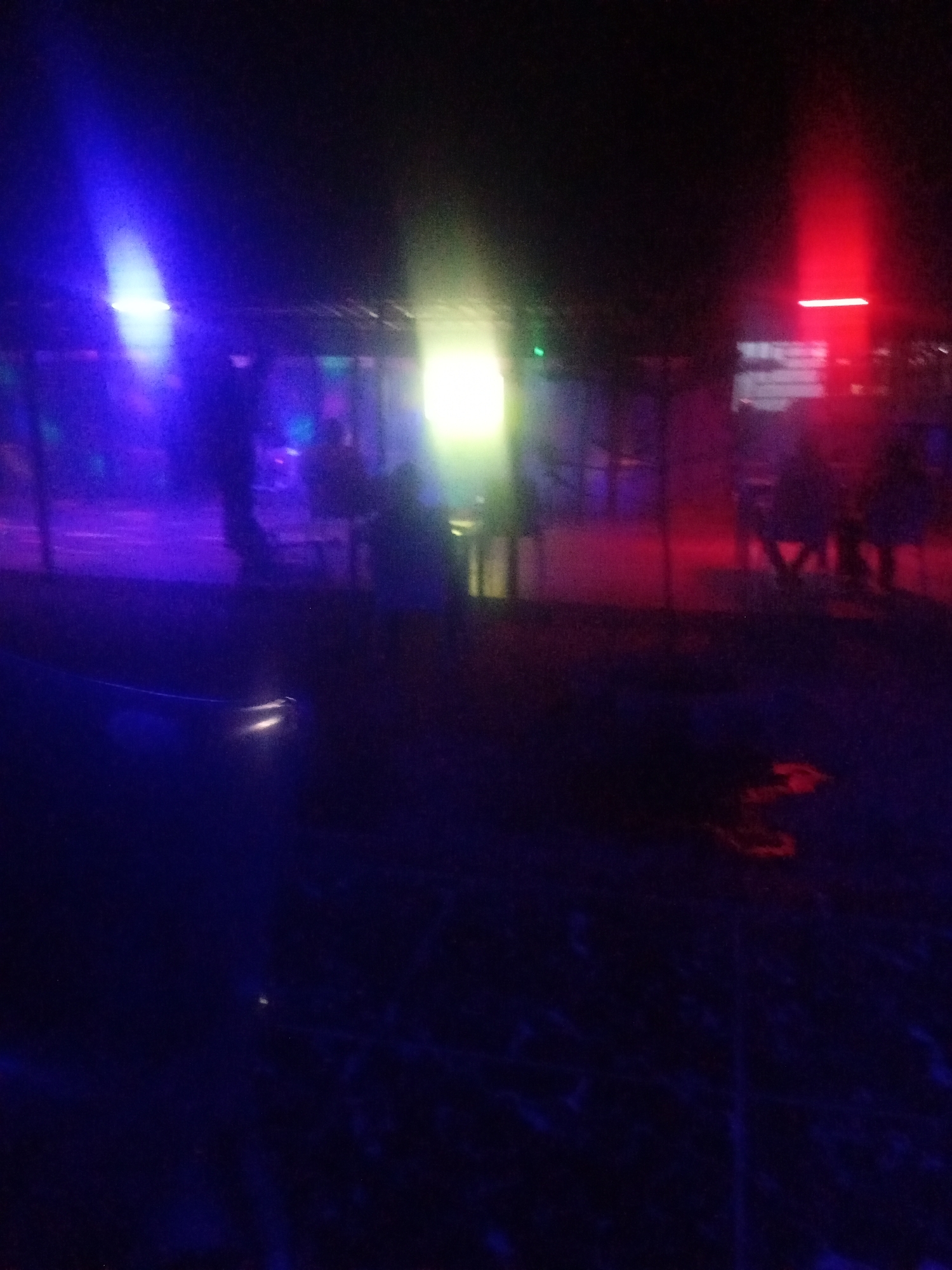 The image shows a dimly lit outdoor area at night, illuminated by various colored lights, including blue, green, and red. There are silhouetted figures of people in the background, some appearing to be seated at tables. The overall scene suggests an outdoor gathering or event, possibly a festival or a party, with a somewhat blurred and atmospheric quality due to the low light conditions and the use of colored lighting. The ground appears to be dark and possibly uneven, with some scattered debris.