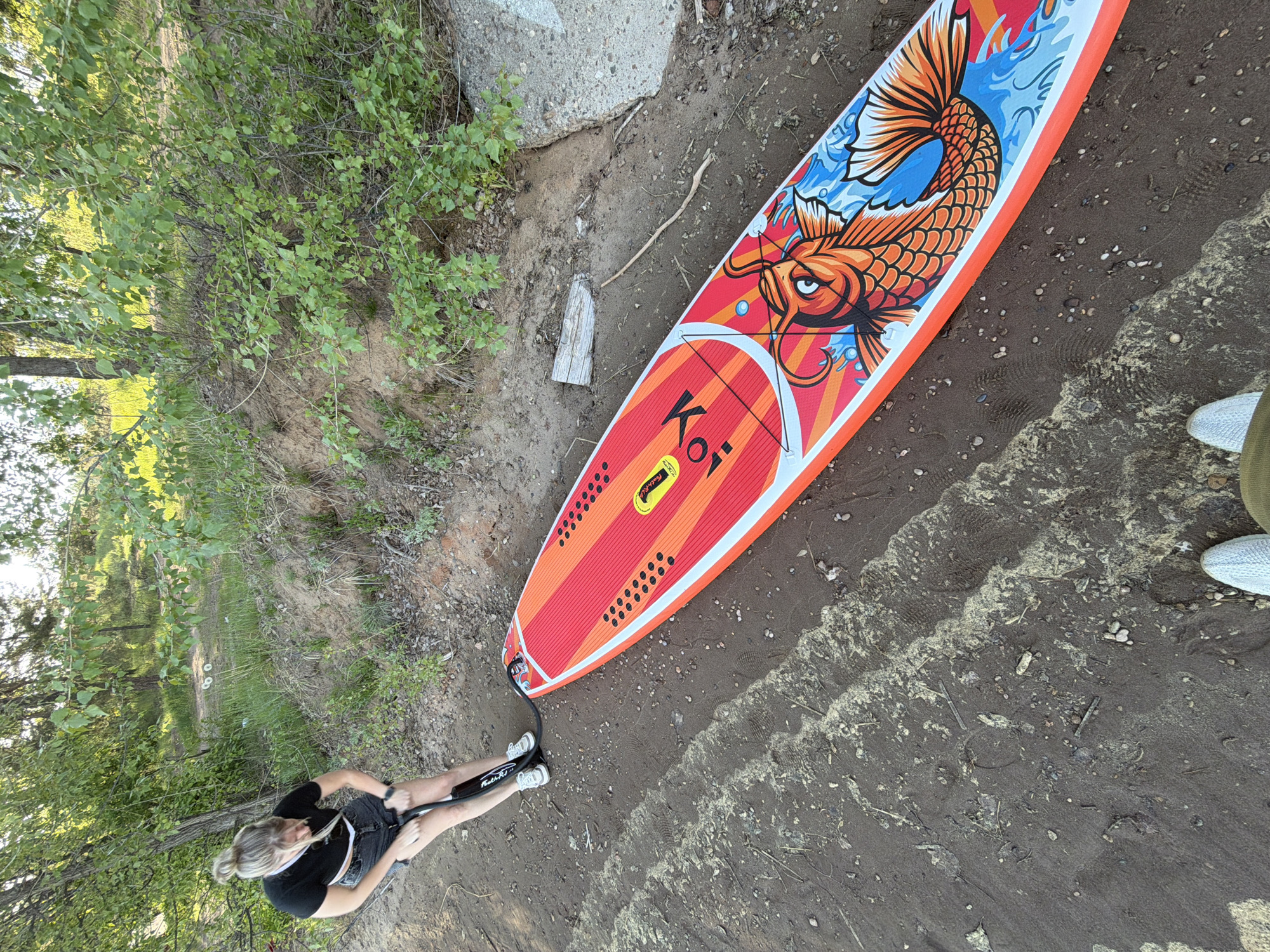 The image depicts a woman inflating a "Koi"-branded paddleboard on a muddy shore, likely near a body of water. The paddleboard is red, orange, and white, adorned with a Koi fish design. The woman wears a black top and denim shorts, and white shoes. She appears to be using a hand pump connected to the board. The background shows foliage and trees, along with what could be a concrete structure. The location is the city of Chaikovsky, Russia.