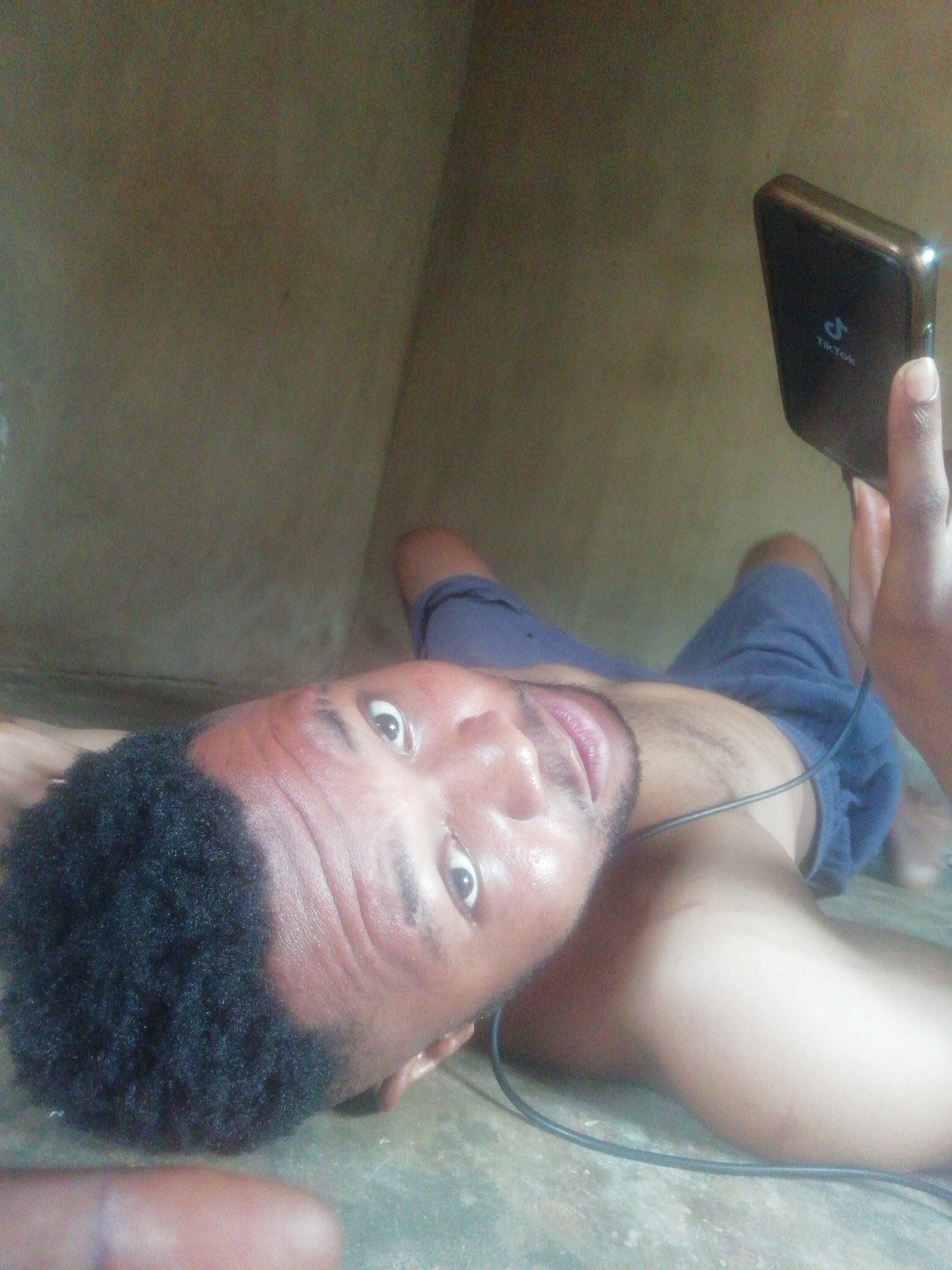 A shirtless man is lying on what appears to be a dirty floor in a room with drab, poorly lit walls. He is holding a black mobile phone with the TikTok logo on the back. An audio cable is draped across his neck. His expression is a startled gaze directly at the camera. The location is unknown.