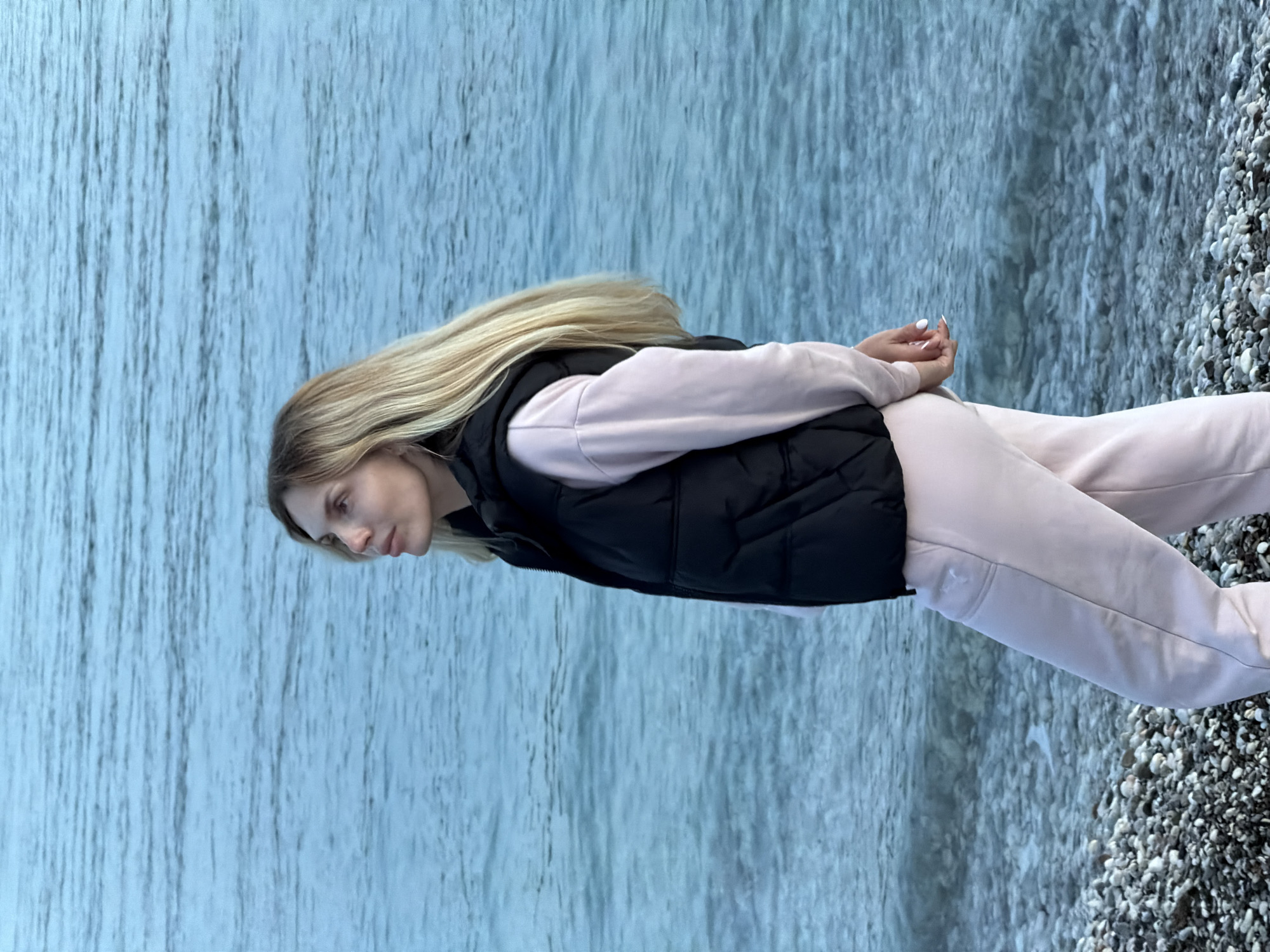 A light-skinned woman with long blonde hair stands with her back toward the camera on a pebble beach. She wears a light pink sweatsuit with a black puffer vest. Her hands are clasped behind her back. In the background is the Adriatic Sea. The scene appears to be in Budva, Montenegro, based on the rocky beach and Adriatic Sea.