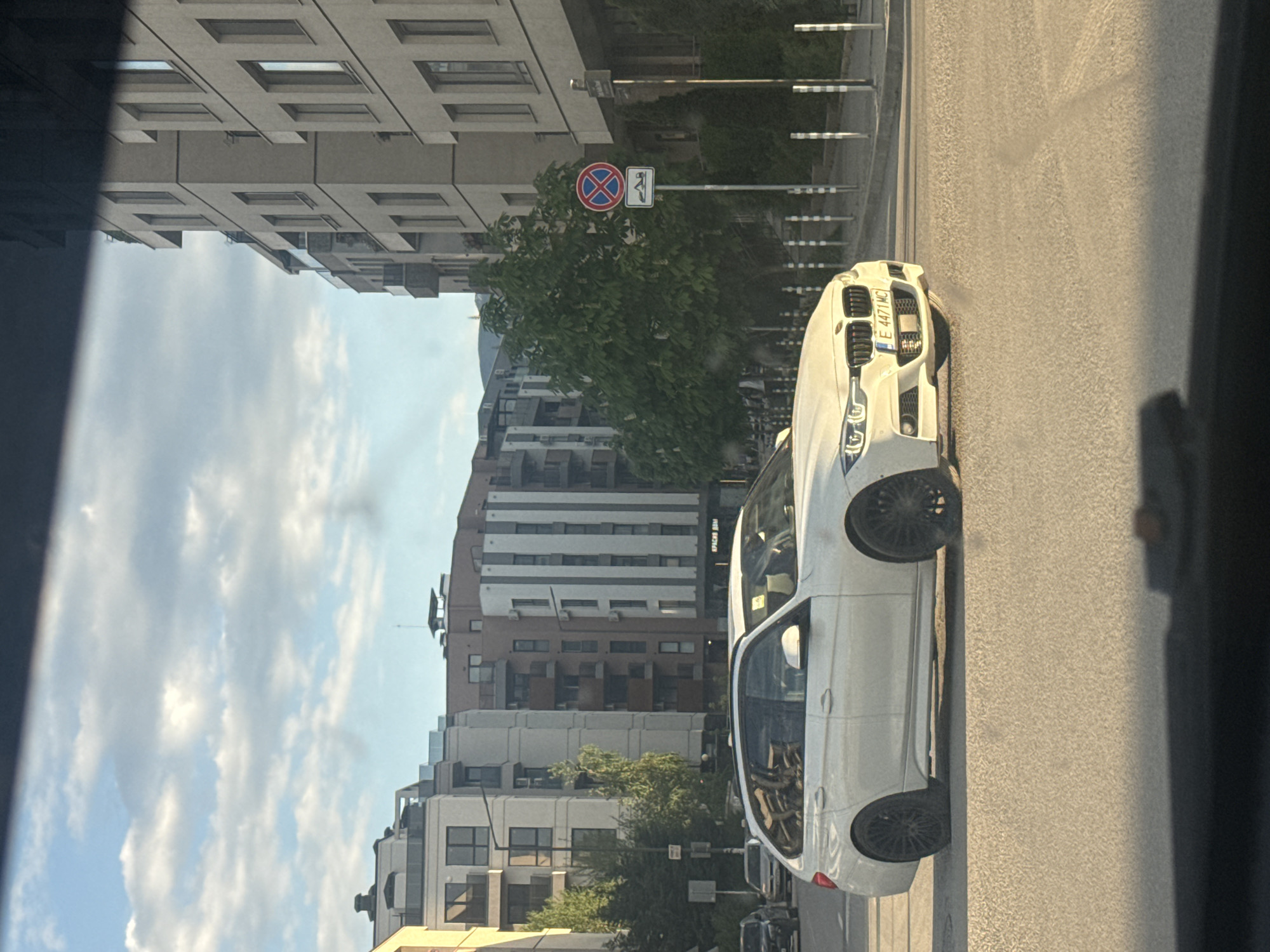 The image shows a street scene in Sofia, Bulgaria. A white car is parked on the right side of the road, with its front facing the camera. The license plate number is "E 4471 MC." A "no parking" sign is visible on a pole behind the car, near a tree. Buildings line the street, with a mix of residential and commercial structures. The sky is visible with some clouds. The image appears to be taken from inside another vehicle, looking out onto the street.
