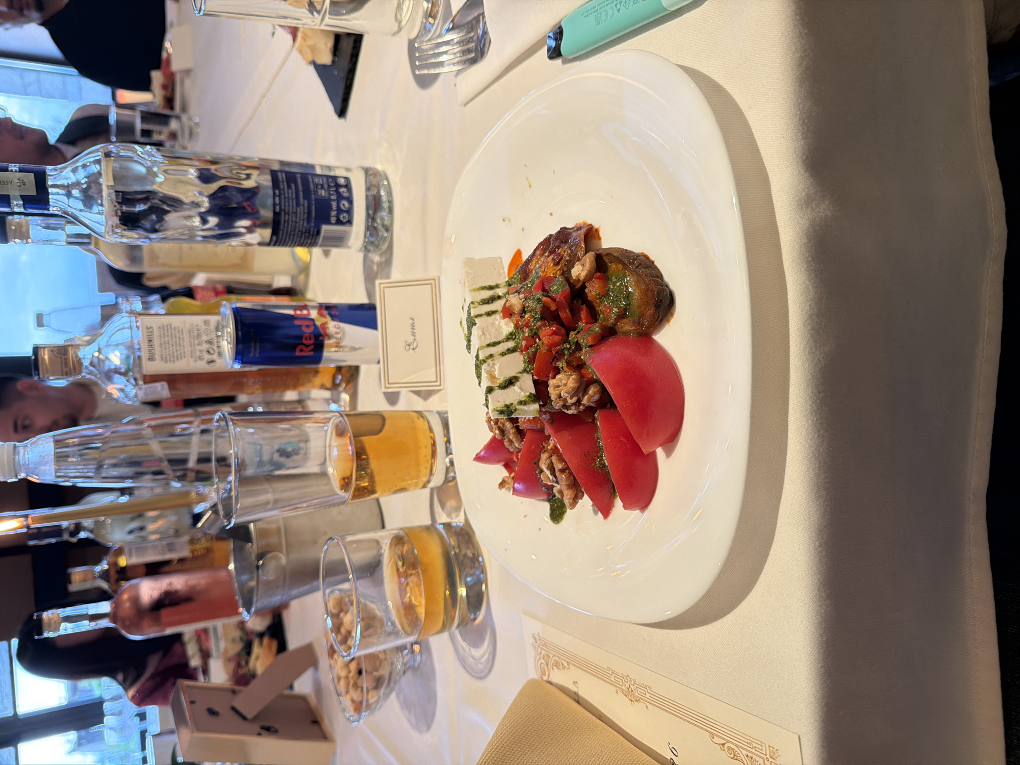 The image shows a table setting with various items. A white plate is in focus, featuring a salad with tomatoes, eggplant, and cheese drizzled with pesto. Nearby, there are several bottles of alcohol, including vodka and Red Bull, along with glasses, some filled with liquid. A place card with the name "Emma" is also visible. The table is covered with a white tablecloth. The scene takes place indoors, likely at a restaurant or catered event. I have no information about the location in the city of Sofia, Bulgaria.