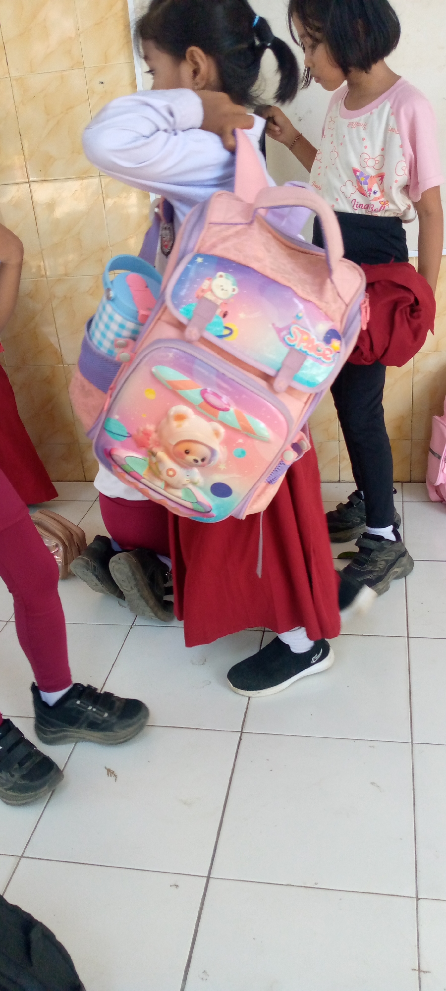 In Pacing, Indonesia, a group of young girls are gathered indoors, possibly in a school or community center, as indicated by the tiled floor and light-colored tiled wall. The central focus is a girl wearing a light purple long-sleeved shirt and a maroon skirt, carrying a large pink and purple backpack adorned with cartoon bears and space imagery. Another girl to her right wears a pink and white t-shirt with cartoon cat graphics and black pants. A third girl, partially visible on the left, wears maroon leggings and a white long-sleeved shirt. The girls appear to be interacting, with one girl adjusting the strap of the backpack. Shoes visible on the floor include black sneakers with velcro straps and a pair of black slip-on shoes. The overall scene depicts a casual, everyday moment among children.