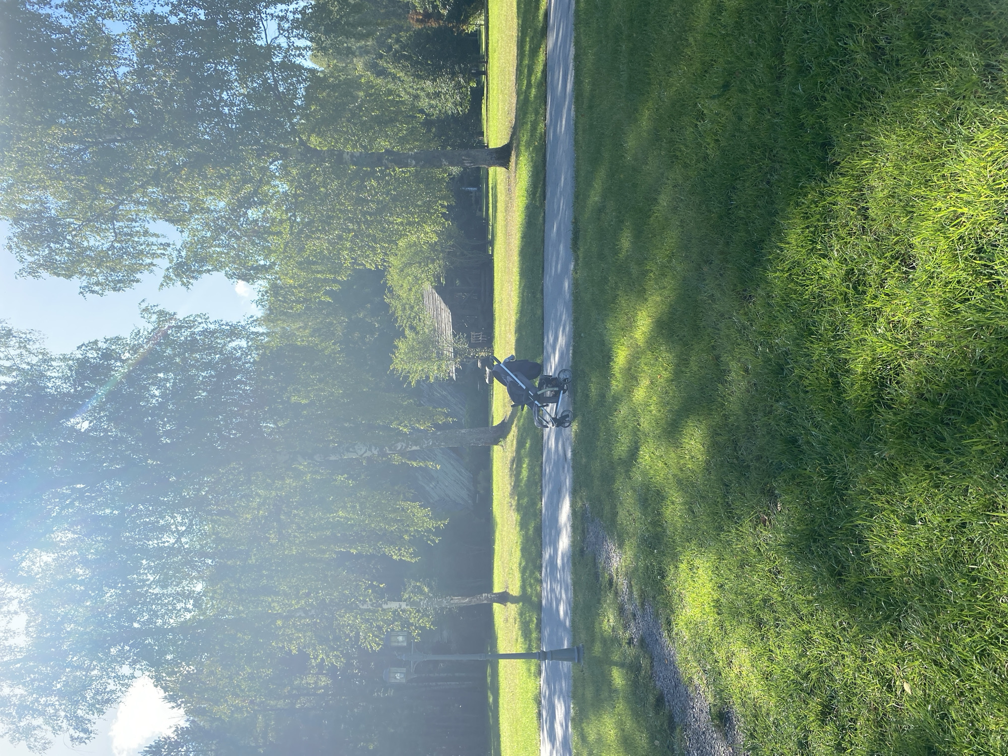 A person is pushing a stroller on a paved path in a park. The sun is shining brightly through the trees, casting shadows on the grass. There is a lamppost on the left side of the image, and a wooden building in the background. The location is the Chulis Museum of Folk Architecture and Rural Life in Chulis, Russia.