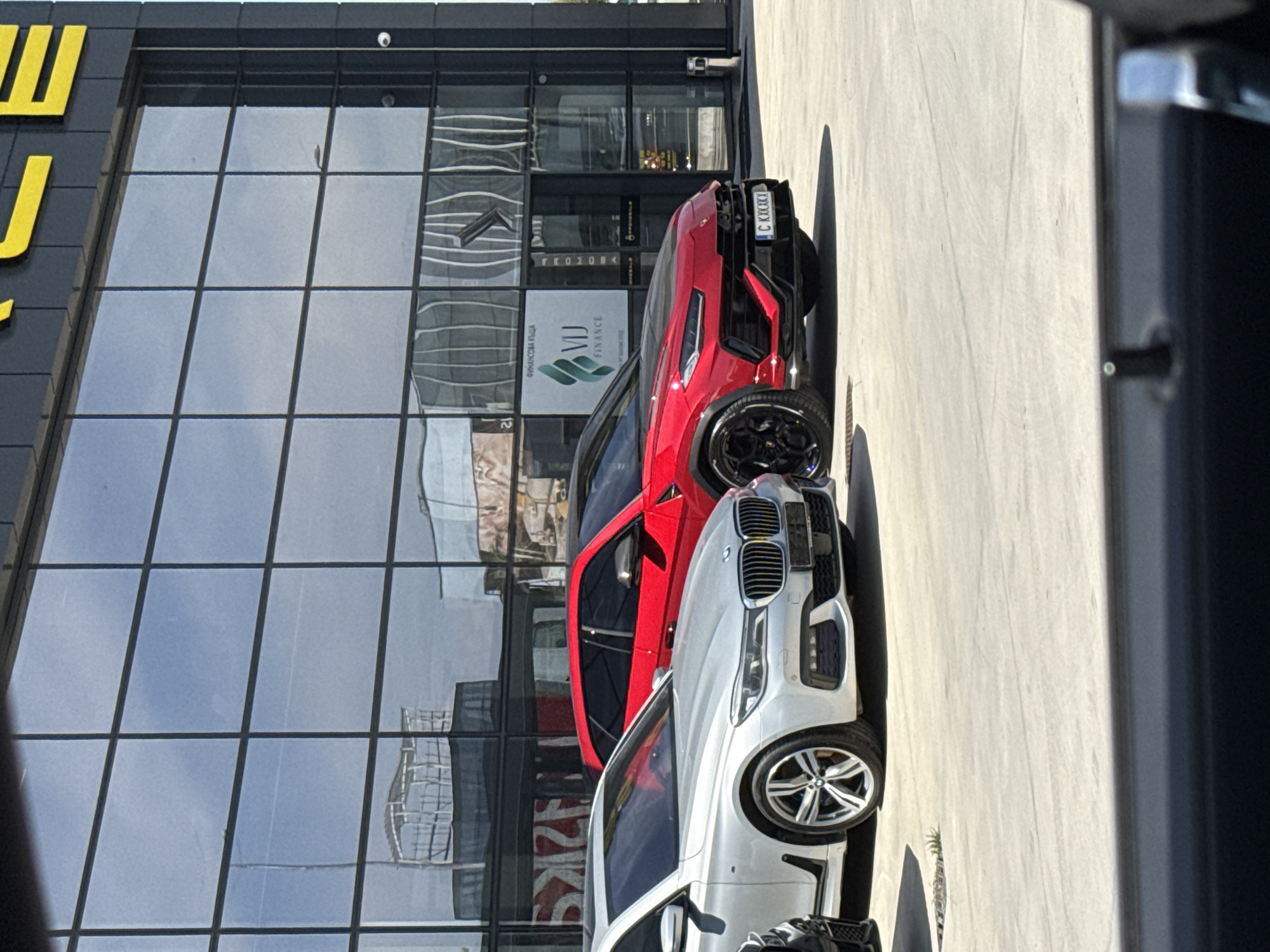 This image, when viewed upright, displays a sunny daytime scene in Plovdiv, Bulgaria, featuring two luxury SUVs parked in front of a modern commercial building. The building on the left has a dark grey facade with large, gridded glass windows that reflect the bright sky and distant structures. A small white security camera is mounted on the upper part of the facade. Partially visible yellow Cyrillic letters on the building's exterior suggest a business name.
Two high-end vehicles are parked parallel to the building. In the foreground is a silver or white BMW SUV, likely an X5 or X6, identifiable by its distinctive front grille and multi-spoke wheels. Behind it is a vibrant red Lamborghini Urus SUV, showcasing its bold rear design, black multi-spoke wheels, and a personalized Bulgarian license plate that reads "C KKK KXX". The "C" prefix on the plate indicates registration in Sofia province.
Through the reflective glass windows of the building, a white sign is clearly visible, displaying a green wave-like logo and the text "VJ FINANCE". Below this, in Cyrillic, it reads "ФИНАНСОВА КЪЩА", which translates to "Financial House". The ground where the vehicles are parked is a light-colored, paved surface, brightly illuminated by direct sunlight, casting sharp shadows from the cars and the building. The scene depicts a commercial setting on a clear and sunny day.