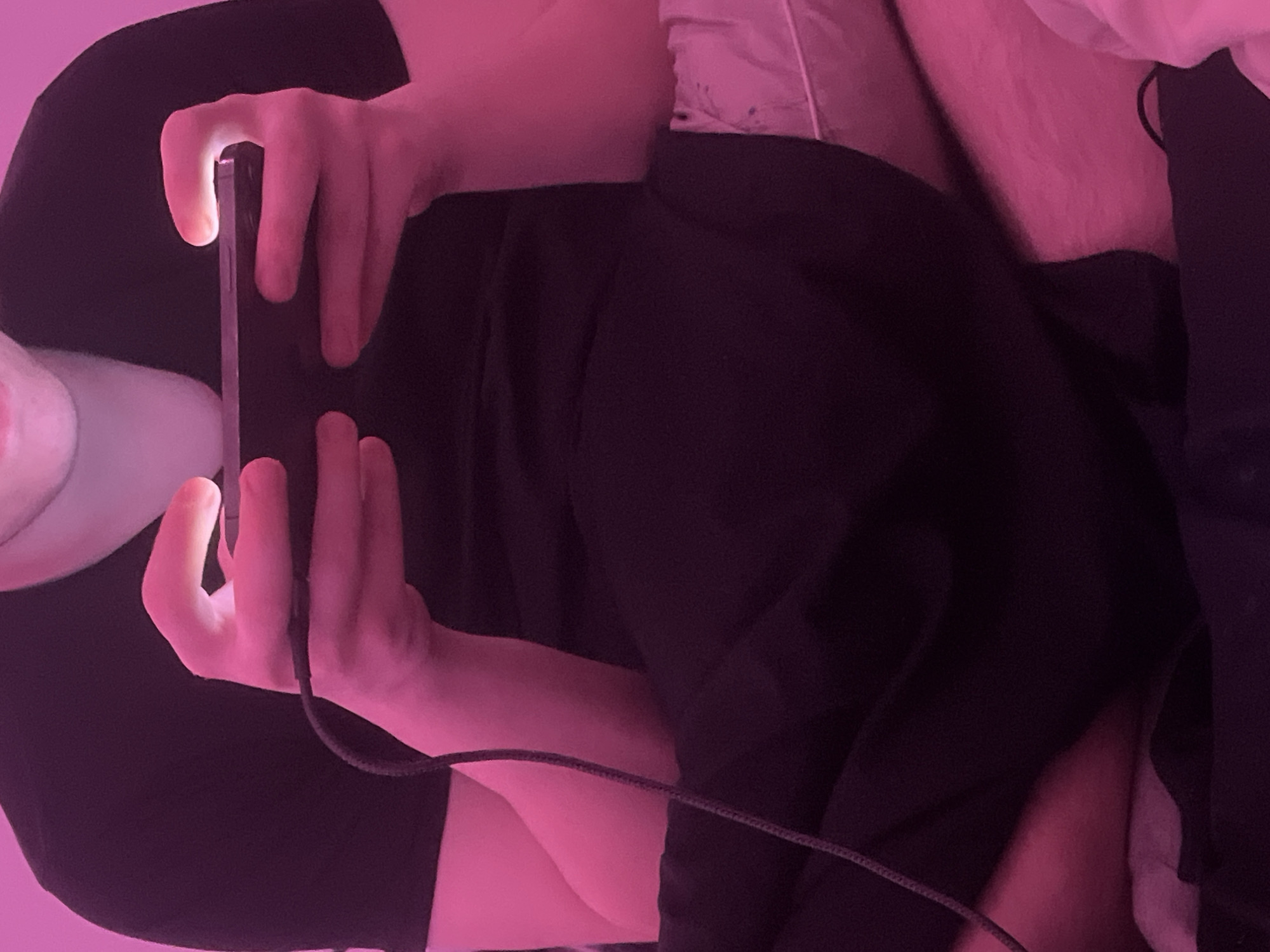 A person is reclined on a bed, illuminated by a pink light source. They are holding a smartphone horizontally, with both hands gripping the device. The screen of the phone appears brightly lit, suggesting it is in use. A charging cable is attached to the phone. The individual is wearing a dark-colored garment. A pillow is visible behind them. The location in the city of Saint Petersburg, Russia is not evident from the image.
