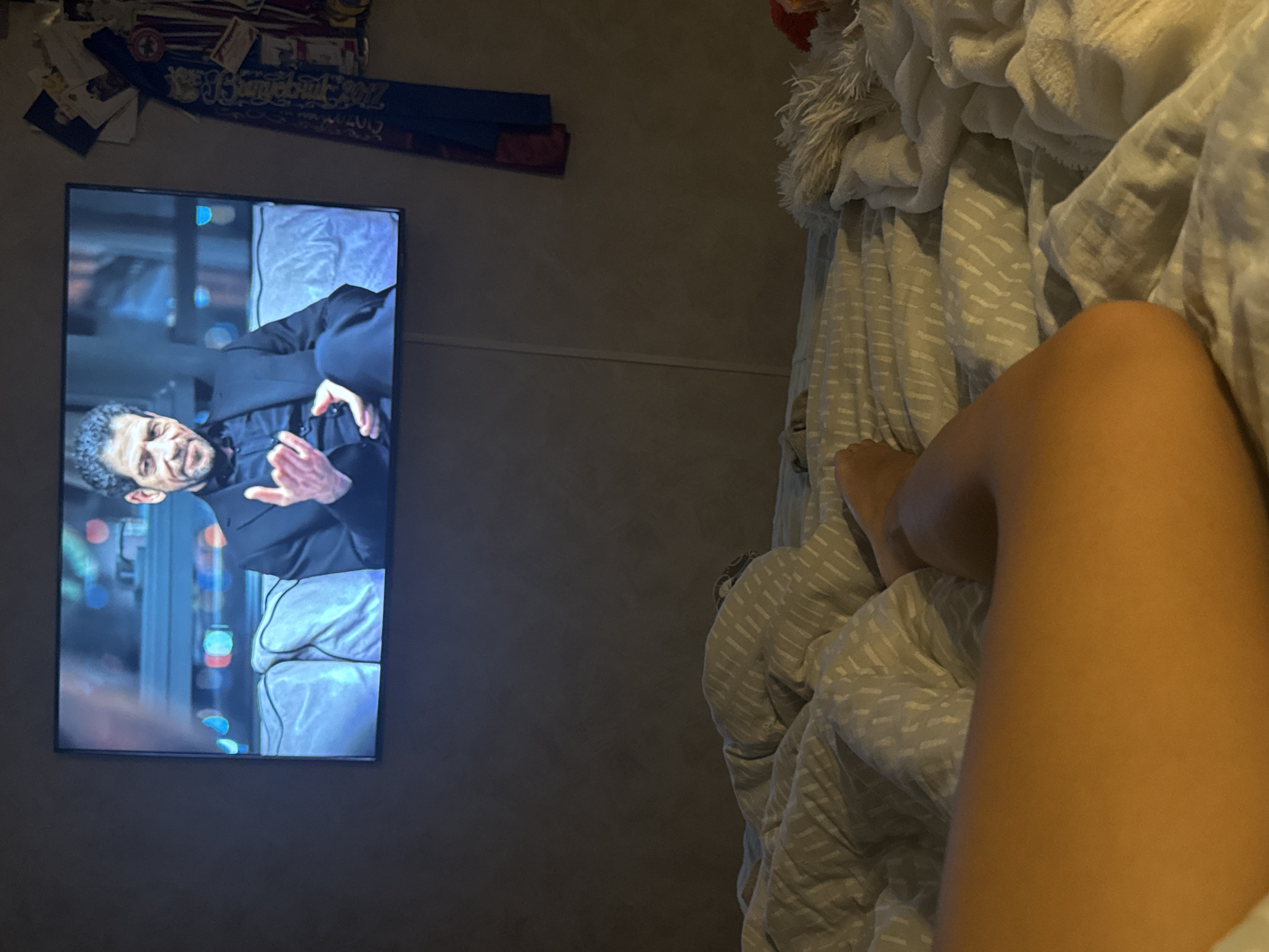 A television displaying a man in a dark suit is positioned on a wall. To the right of the television, a bed with rumpled white and gray patterned linens is visible, with a leg extending from under the covers. An indiscernible collection of items is located above the television. The location in Chaikovsky, Russia, cannot be determined from the image.