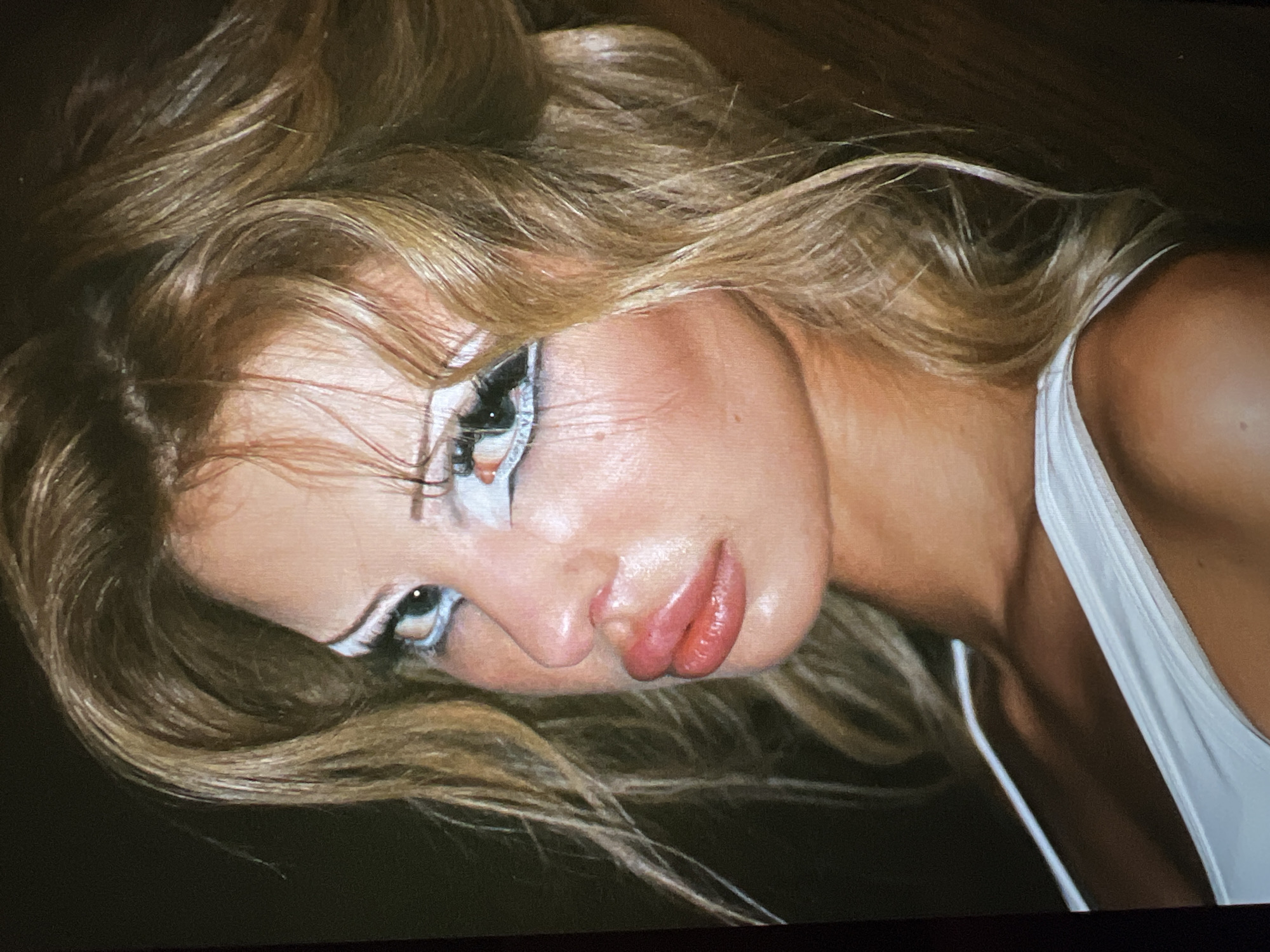 The image is a close-up portrait of a woman with blonde hair, styled in loose waves. She is wearing a white top. Her face is heavily made up with dramatic white eyeliner and visible eyelashes. Her lips are full and appear to be glossed. The background is dark and nondescript. There is no visible indication of the city of Saint Petersburg, Russia, or any specific location.