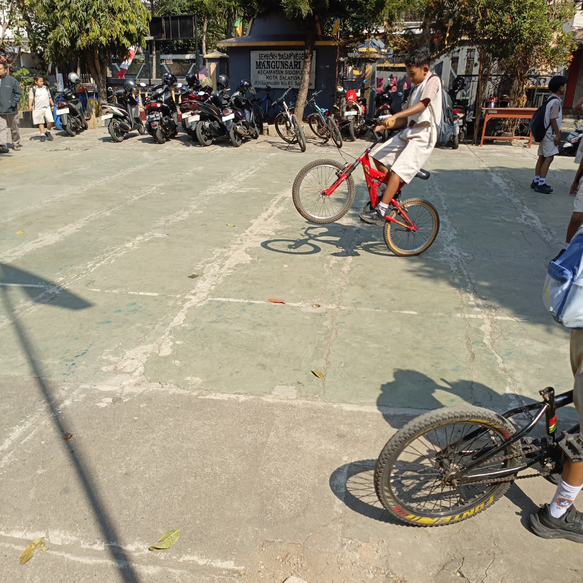 The image depicts an outdoor scene in Salatiga, Indonesia, likely at a school, indicated by the sign "SEKOLAH DASAR NEGERI MANGUNSARI" which translates to "MANGUNSARI STATE ELEMENTARY SCHOOL". The foreground shows a young boy on a red bicycle, performing a wheelie. He is wearing a school uniform consisting of a white shirt with checkered sleeves and light-colored shorts, white socks, and dark shoes. His bicycle is a BMX-style bike with a red frame and knobby tires.
In the background, a line of parked motorcycles is visible, suggesting this area serves as a parking lot or is adjacent to one. Another young boy, also in school uniform, stands to the right, facing away from the camera. He is wearing a backpack. Other individuals, presumably students and perhaps a teacher, are present in the background, some in school uniforms. The environment is a paved outdoor area, likely a courtyard or sports court, with some trees and a building with signage. The lighting indicates it is daytime with bright sunlight, casting distinct shadows. Another bicycle is visible in the lower right portion of the image, its rear wheel and pedals clearly in view.