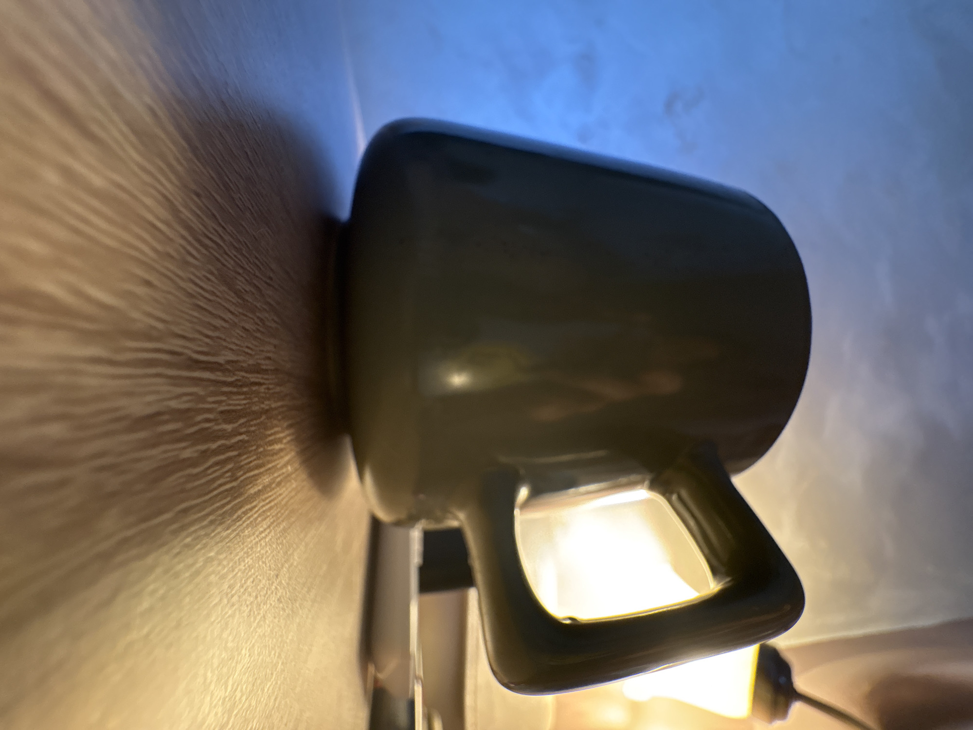 This image, captured in Bar, Montenegro, presents a close-up, low-angle view of a dark-colored, likely grey or dark green, ceramic mug. The mug is the central subject, dramatically illuminated by a strong, warm-toned artificial light source positioned beneath and slightly behind it. This light casts bright highlights on the mug's handle and the lower part of its body, creating a high-contrast scene.
The mug rests on a textured wooden surface, which could be a table or countertop. The intense light emphasizes the natural wood grain, creating distinct, linear patterns of light and shadow that stretch across the surface, receding towards the left side of the frame. In the background, above and behind the mug, a light blue surface is visible, contrasting with the warm illumination below. This blue area suggests a wall or ceiling.
The scene appears to be an indoor setting, characterized by artificial lighting, indicating it might be evening, night, or a room with limited natural light. No people or specific activities are depicted; the image focuses solely on the still life arrangement. There is no visible text. The composition highlights the interplay of light, shadow, and texture.