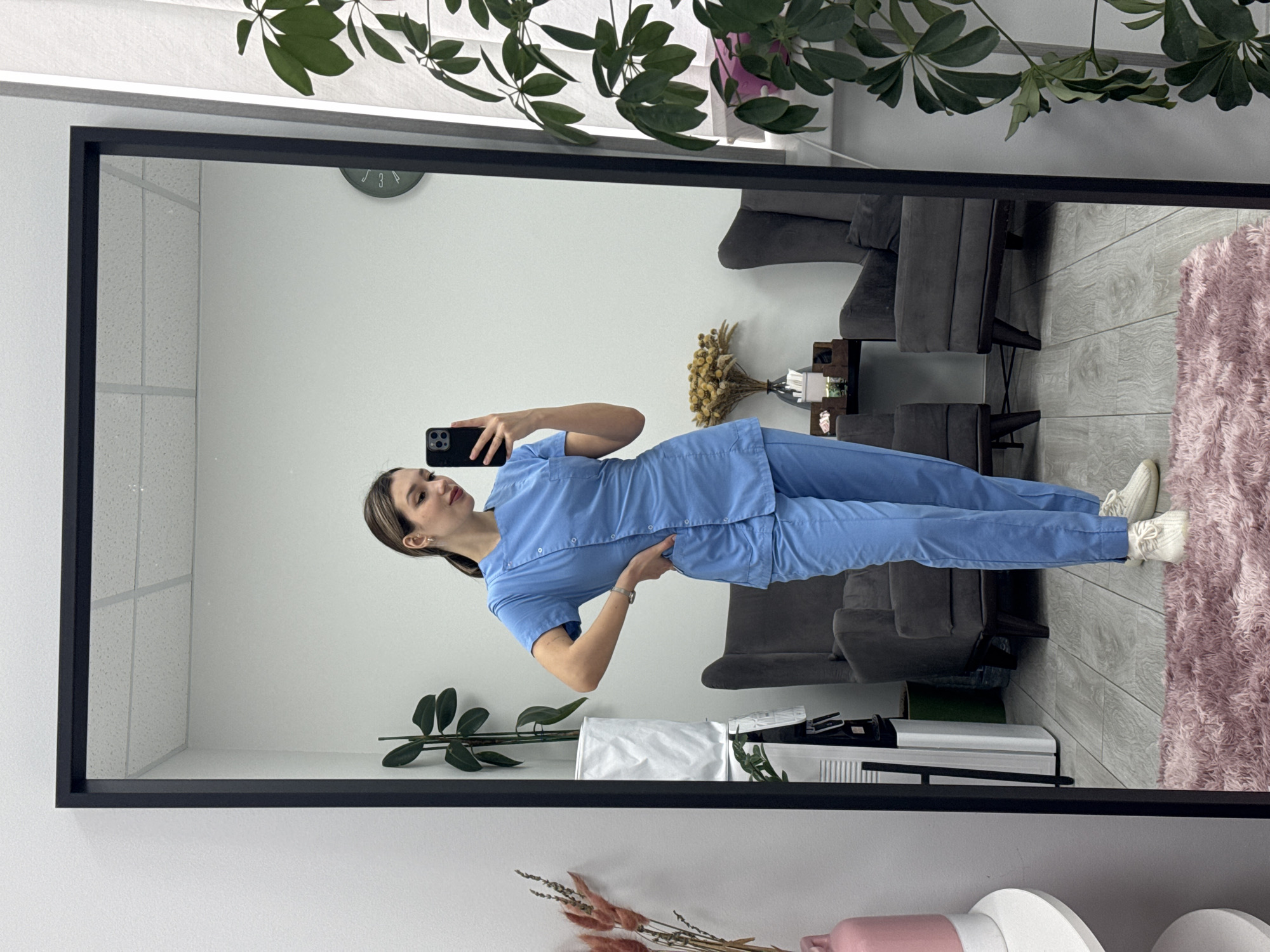 A full-shot image, rotated 90 degrees clockwise, captures a woman taking a selfie in a large mirror. The woman stands upright within the mirror's reflection, holding a dark-colored smartphone to capture her image. She is dressed in light blue medical scrubs, consisting of a short-sleeved top with buttons and matching pants, paired with white sneakers. A silver-toned watch is visible on her left wrist, and she wears small earrings. Her right hand is placed on her hip as she poses, looking towards her phone screen with a slight smile.
The mirror itself has a thick black frame. The reflected room behind the woman appears to be an indoor setting, possibly a waiting area, office, or clinic. Two dark grey upholstered armchairs are visible in the reflection, arranged with a small dark wooden side table between them. On the table, there's a small bouquet of dried yellow flowers and some white items, possibly tissues or paper. The floor is light-colored wood or laminate, and a fluffy pink rug partially covers it on the right side of the reflection. The walls are light-colored, likely white. High on the reflected wall, a green-faced round clock shows partial numbers "3" and "4". A large, leafy green plant is reflected in the upper part of the mirror, and another smaller plant with dark green leaves is reflected on a lower surface to the left. The ceiling appears to have a grid pattern, indicative of suspended ceiling tiles.
Outside the mirror's frame, at the top, a vibrant green plant with large leaves hangs over, and at the bottom, a pink vase with dried pinkish-brown flowers and some white decorative objects are visible. The overall lighting suggests an indoor setting, likely during the daytime.