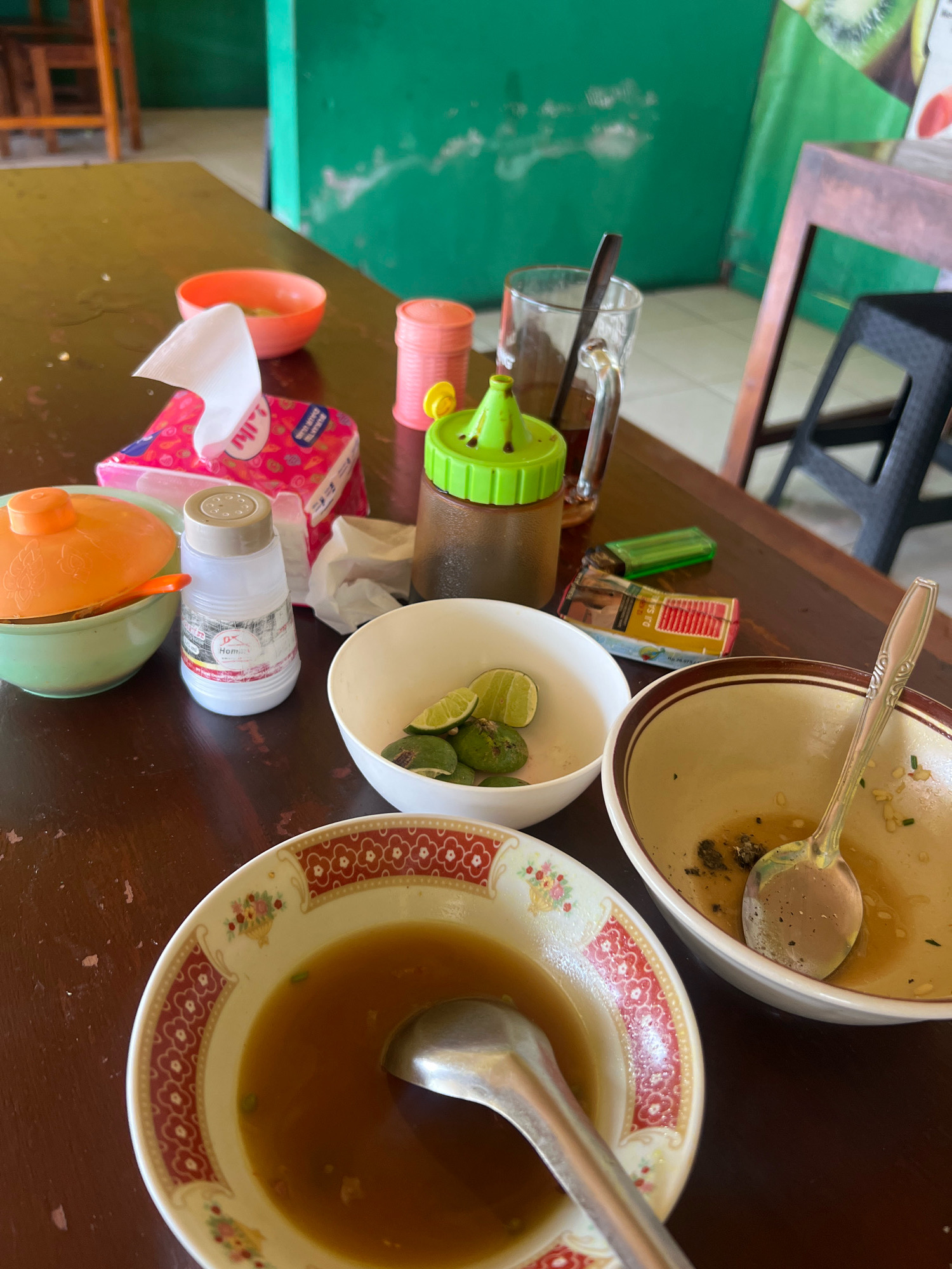 A close-up, overhead shot captures a wooden table laden with remnants of a meal. In the foreground, a bowl with a decorative red and white rim contains a light brown broth, with a spoon resting inside. To its right, another bowl, nearly empty, holds a spoon and scattered food debris. To the left, a green bowl with an orange lid sits beside a light-colored shaker, possibly for salt or pepper, labeled "DX Home". In the background, a pack of tissues with a pink wrapper is visible, alongside a glass of iced tea with a spoon. A green-capped condiment bottle and a lighter are also present, along with a pile of lime wedges in a white bowl. The scene appears to be set in an outdoor dining area, suggested by the green wall and natural light. The location is identified as Sayegan, Indonesia.