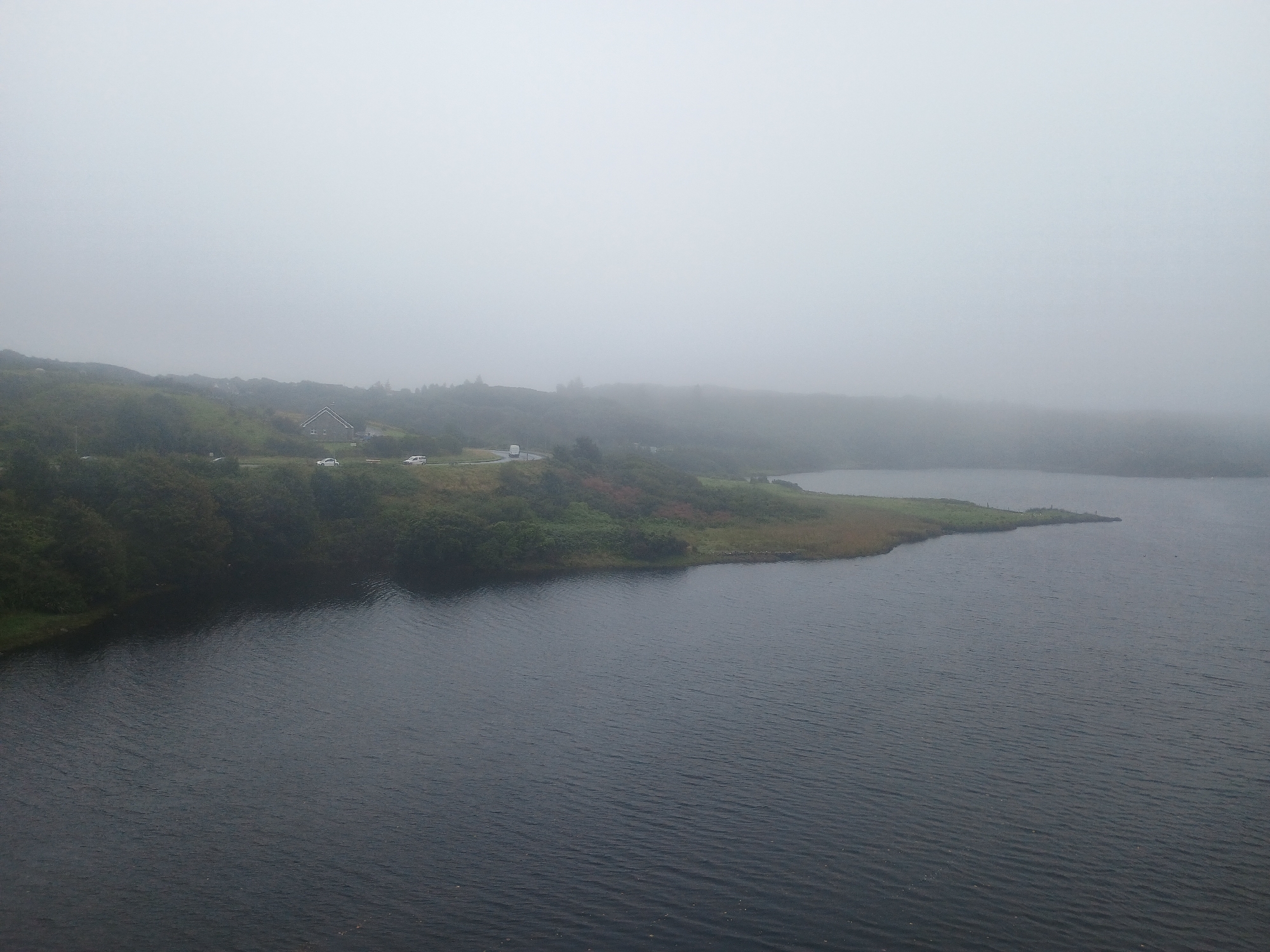 This is an aerial drone photograph of a vast, dark body of water adjacent to a landmass covered in vegetation and mist. The water occupies the majority of the foreground and extends towards the right side of the frame. The landmass, on the left and upper portions of the image, is characterized by rolling hills with green and brown patches, indicative of grass and possibly some trees or shrubbery. A road with a few vehicles is visible winding along the edge of the land. The background is dominated by a dense, uniform white mist that obscures any distant features, creating a hazy and ethereal atmosphere. The sky is indistinguishable from the mist.
