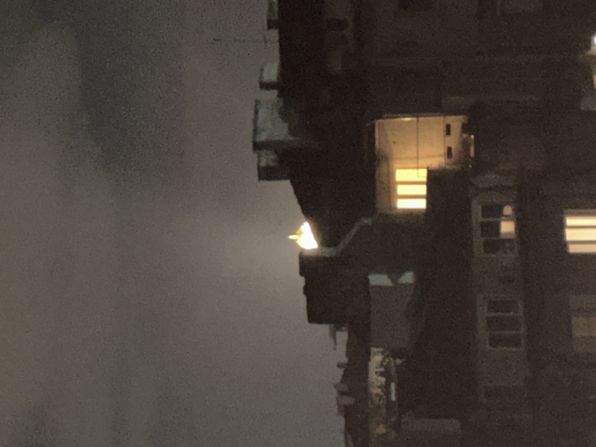 The image shows a night scene of a building in what might be Sofia, Bulgaria, though without more context, definite confirmation is impossible. A tall, dark building is on the right with visible windows and some lit-up rooms. The left side of the image is darker, potentially showing a cloudy sky or a shadowed area. A light is visible on the upper part of the building. The overall impression is a dark, urban night setting.