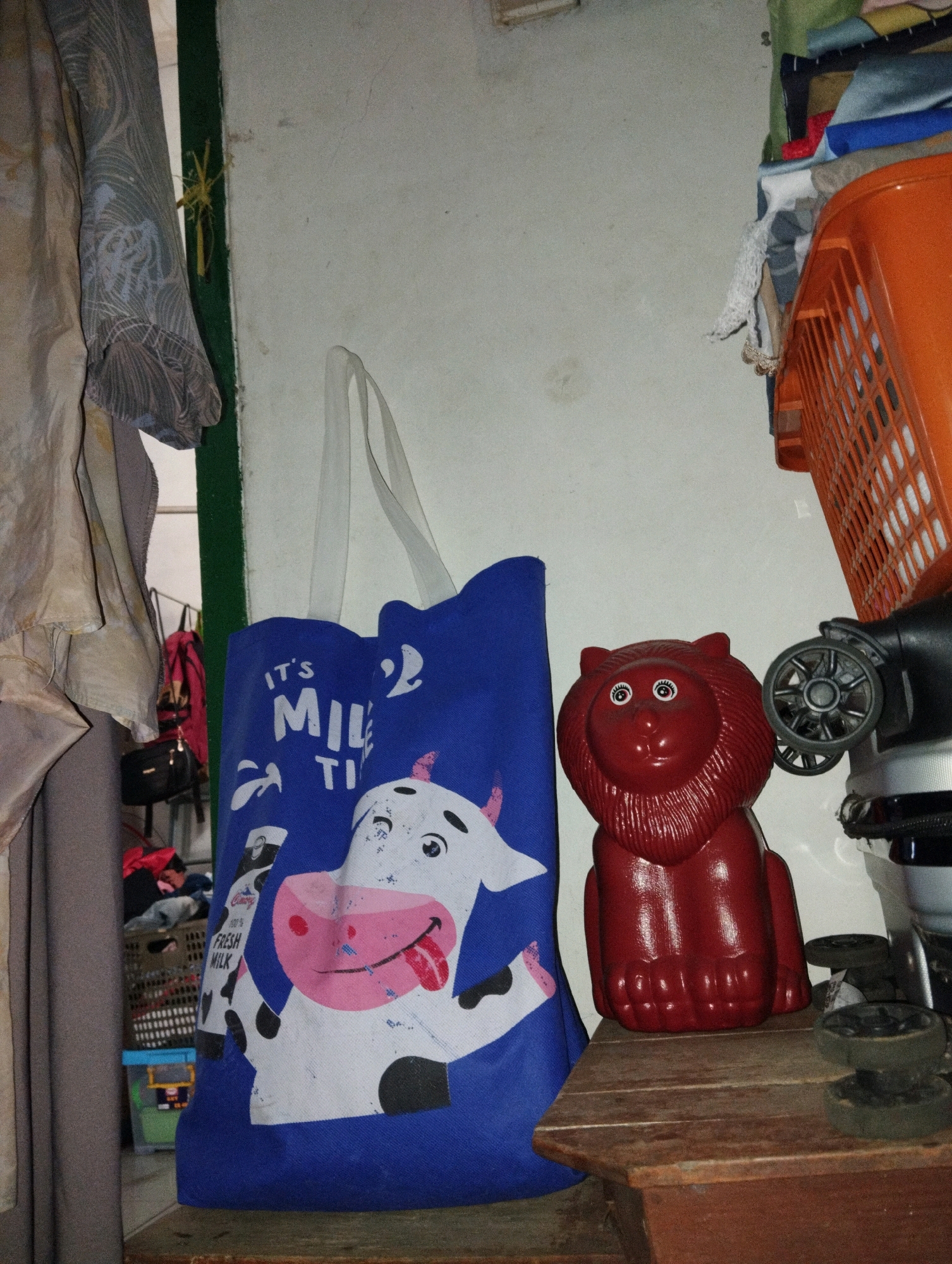 In what appears to be a cluttered storage area or a small room in Tiparraya, Indonesia, a blue tote bag with a graphic of a cow and the text "IT'S MILK TIME" is prominently displayed. To the right of the bag, a shiny red, lion-shaped piggy bank sits on a rustic wooden shelf. Behind the piggy bank, part of an orange plastic basket, a black electronic device with wheels, and some assorted clothing items are visible. The background is a simple wall, and the scene is illuminated by artificial light, casting shadows. To the left of the tote bag, a grey garment hangs, obscuring some of the background. The overall impression is one of domestic clutter and everyday objects.