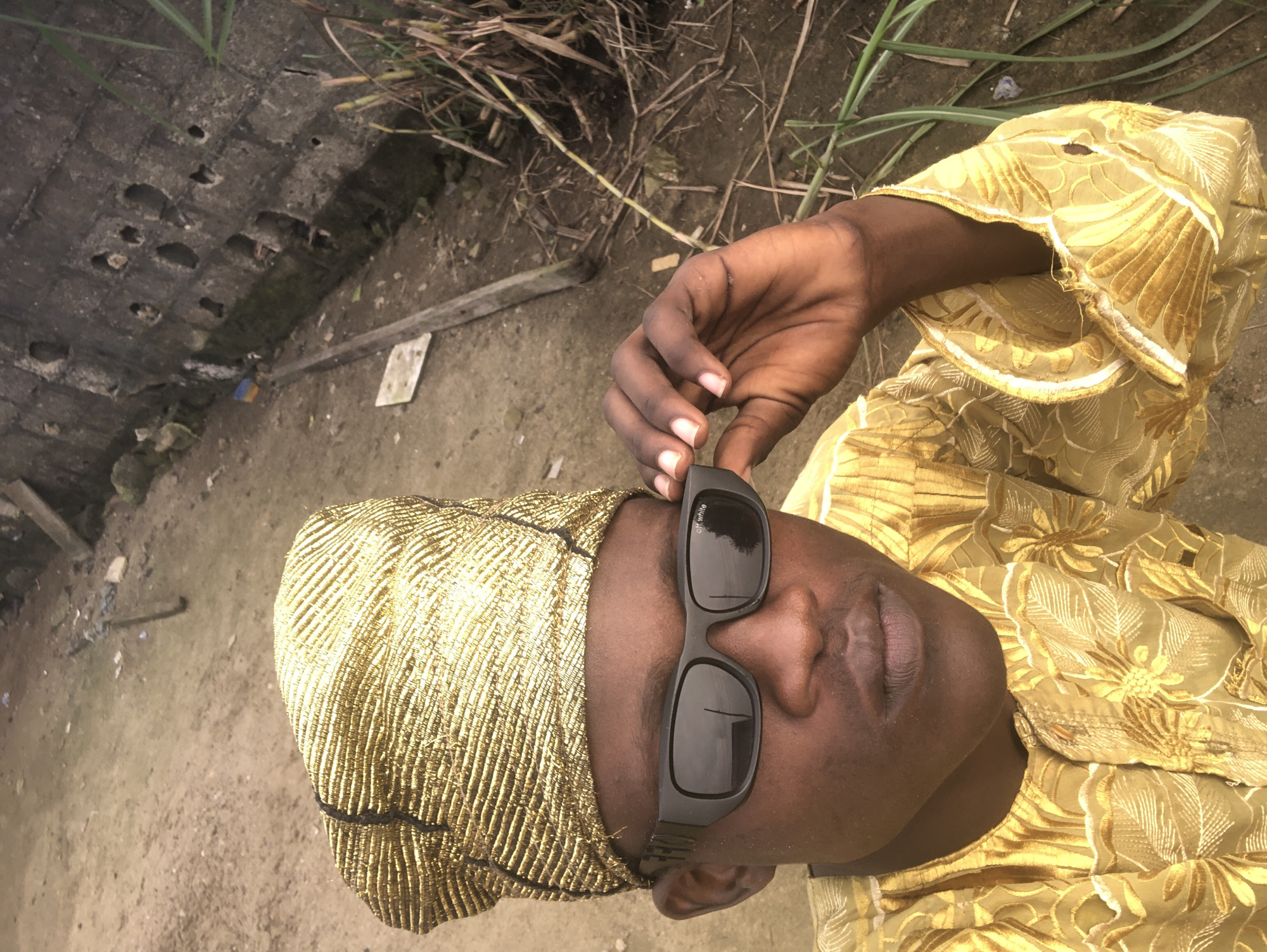 A young Black male is pictured outdoors, in what appears to be a semi-urban or rural area of Ado, Nigeria. He is wearing a gold, intricately patterned traditional Nigerian outfit, known as an Agbada, and a matching gold, textured Fila (headtie). He has dark skin, brown eyes, and is wearing black sunglasses, which he is adjusting with his right hand. The background reveals a dusty, uneven ground with sparse vegetation and a weathered concrete wall with several holes. There are also some discarded wooden planks and debris visible, suggesting an undeveloped or construction site. The lighting indicates it is daytime, with a slightly overcast sky contributing to the muted colors of the environment.