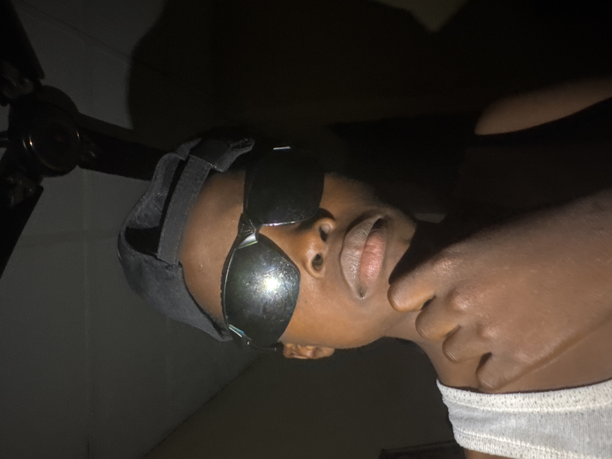 A close-up, low-light photograph depicts a person with dark skin wearing dark sunglasses and a dark head covering, possibly a durag. The person is facing upwards towards the camera. Their right hand is near their neck, with the fingers partially obscuring it. The person is wearing a grey garment around their neck or upper torso. In the background, a ceiling fan is partially visible to the left, and the wall appears to be a light color. The scene is poorly lit. There is no definitive evidence within the image to verify that the location is Onitsha, Nigeria.