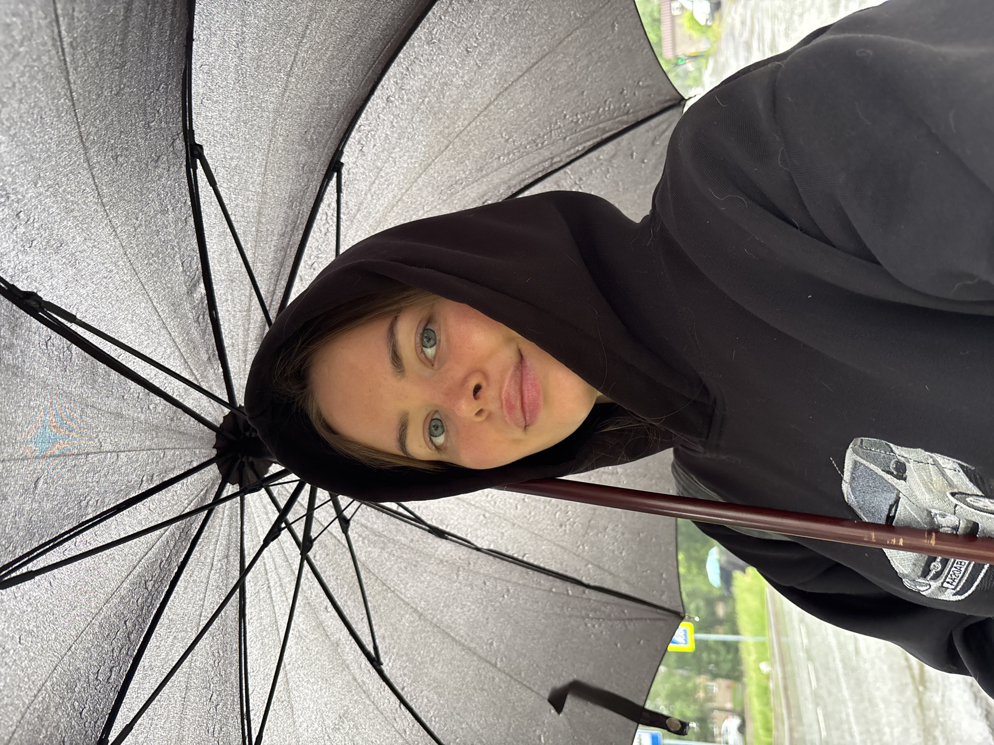 The image captures a young woman from a low angle, holding an open black umbrella in what appears to be rainy weather. The setting is Izhevsk, Russia.
The central subject is a young female with light skin, light-colored eyes (appearing blue or green), and light brown hair, which is partly covered by the hood of her black sweatshirt. She looks towards the viewer with a slight, playful smile, her tongue subtly visible, conveying a lighthearted emotion.
She is holding a large black umbrella overhead, and its underside is clearly visible and speckled with numerous small water droplets, indicating active rain. The black metal ribs and stretchers of the umbrella's frame are distinct. The woman holds the umbrella by its dark brown handle. Her attire includes a black hooded sweatshirt, on which a gray embroidered design of a car is visible, featuring a license plate that appears to read "AA20AB".
The background is blurred, suggesting an outdoor urban or suburban environment. Faint hints of green foliage and a yellow sign or traffic signal can be discerned in the distant, out-of-focus areas. The diffused lighting suggests an overcast, daytime setting. The scene depicts an individual sheltered from rain, capturing a candid and slightly mischievous moment.