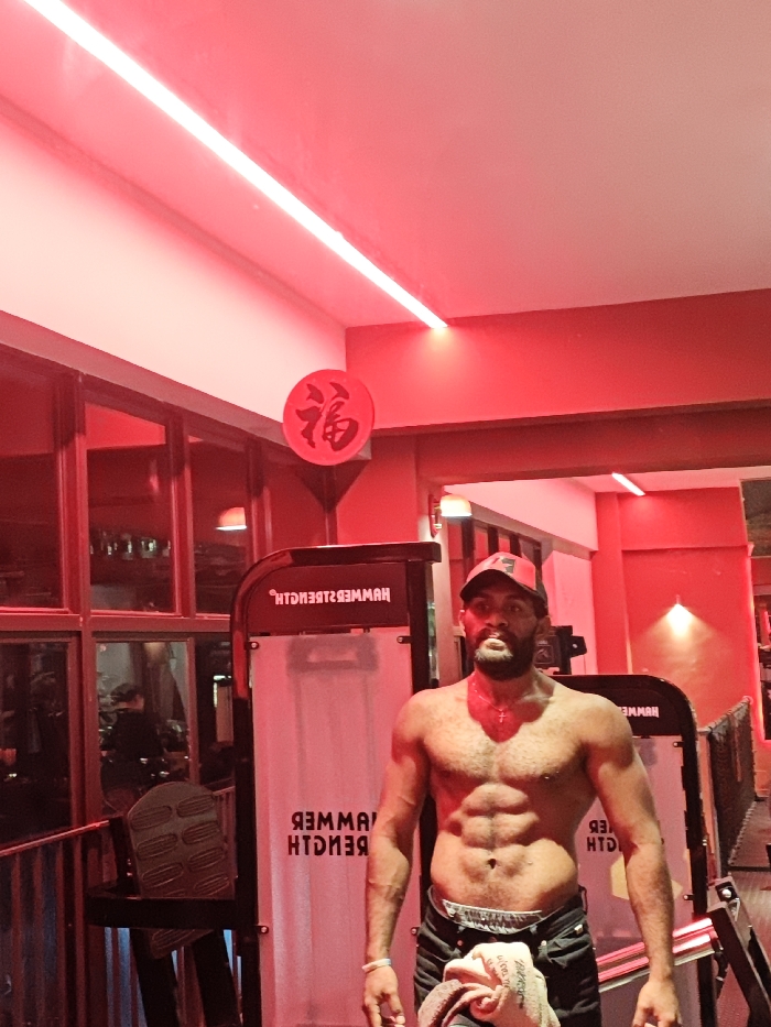 This image captures an adult male posing shirtless within a gym setting in the Chongqing High-tech Zone, China. The environment is characterized by intense red ambient lighting, including a linear ceiling fixture. The subject stands centrally, facing the camera, displaying a muscular torso with defined abdominal muscles. He wears a red baseball cap adorned with a white '7', dark pants or shorts, a white wristband on his left arm, and a small red cross pendant on a chain around his neck. A light-colored towel is tucked into his waistband on the right side. In the background, two pieces of fitness equipment are visible, one directly behind the subject with the reversed brand name 'HAMMER STRENGTH' printed on a white panel. To the upper left, a red circular ornament featuring the Chinese character '福' is mounted on a wall. Large windows are present on the left side, reflecting internal lighting and vague background forms.