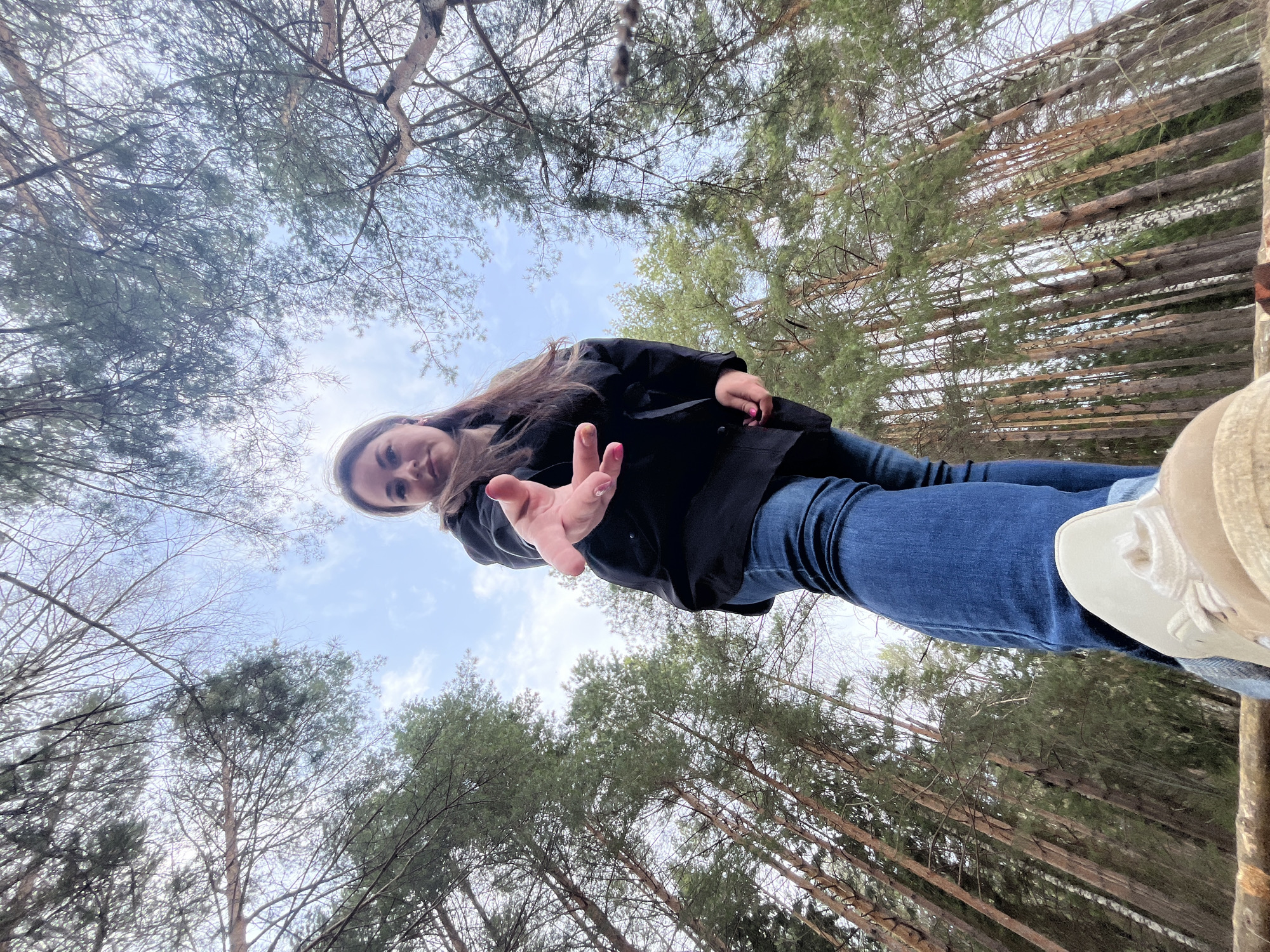 The image is taken from a low angle, looking up towards a young woman standing on a structure made of logs. The woman is wearing a black jacket and blue jeans. She extends her hand towards the camera. The background consists of a forest with tall trees and a partly cloudy sky. The location is Chaikovsky, Russia.