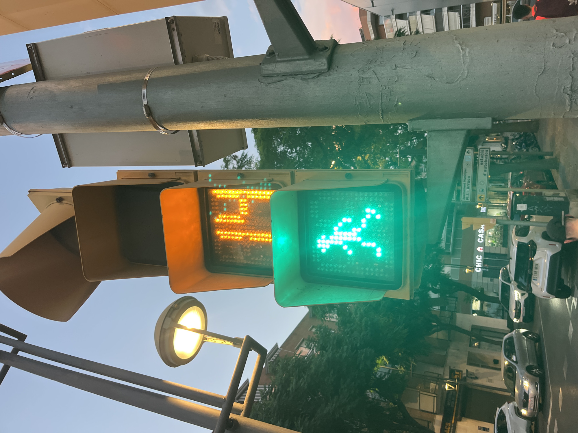 This is a close-up, eye-level shot of a pedestrian traffic light in the city of Tarragona, Spain, likely at an intersection. The traffic light displays a green silhouette of a walking person, indicating it is safe to cross. Above it, the pedestrian signal shows a countdown timer of "14" seconds, displayed in amber LEDs. The traffic light is mounted on a sturdy, grey metal pole, which also supports other electrical equipment. In the background, city buildings and lush green trees are visible, hinting at an urban environment. The sky above shows a twilight hue, suggesting the time of day is either early morning or late evening. The image also captures parts of vehicles on the street, indicating normal traffic activity. There is no direct interaction or action depicted beyond the pedestrian signal indicating the status for crossing.