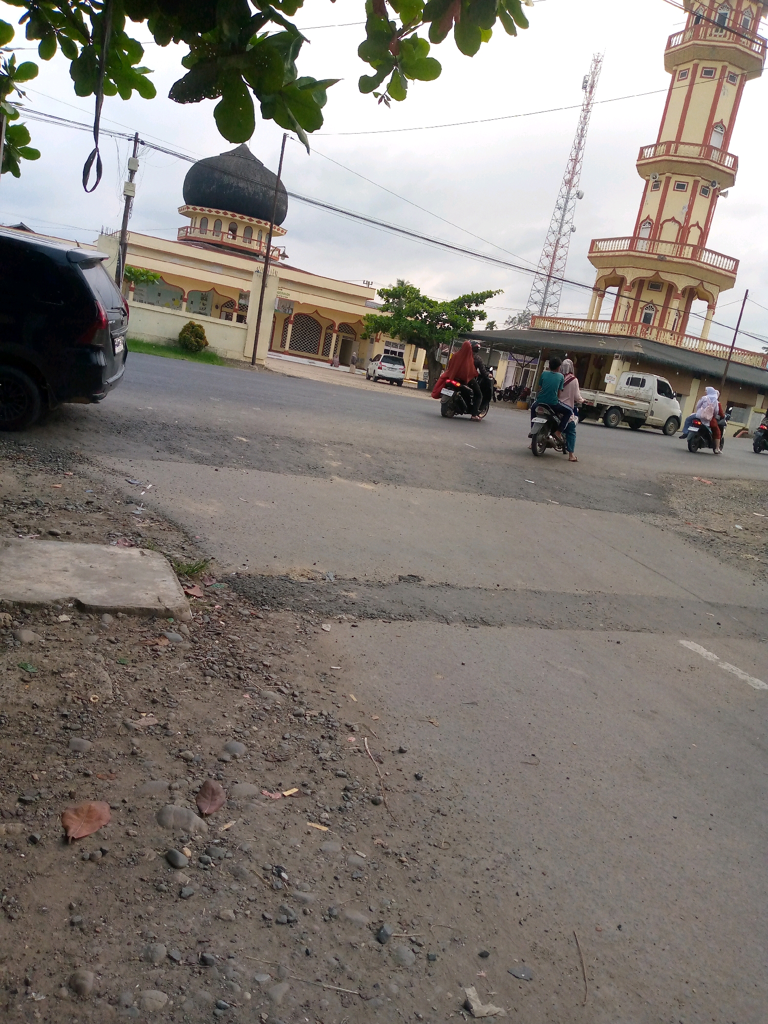 The image shows a street scene in Keudeteupinpunti, Indonesia. A mosque with a dark dome and a tall, cream-colored minaret with red accents are visible in the background. A cellular tower is also present near the minaret. Several people are riding motorcycles on the paved road, with some wearing hijabs. A black car is parked on the left side of the road, and a white truck is also visible. The foreground is dominated by the road surface, which appears somewhat worn, with patches of gravel and dirt. Trees and foliage are present on the left side of the image, partially obscuring the view of the mosque. The sky is overcast.