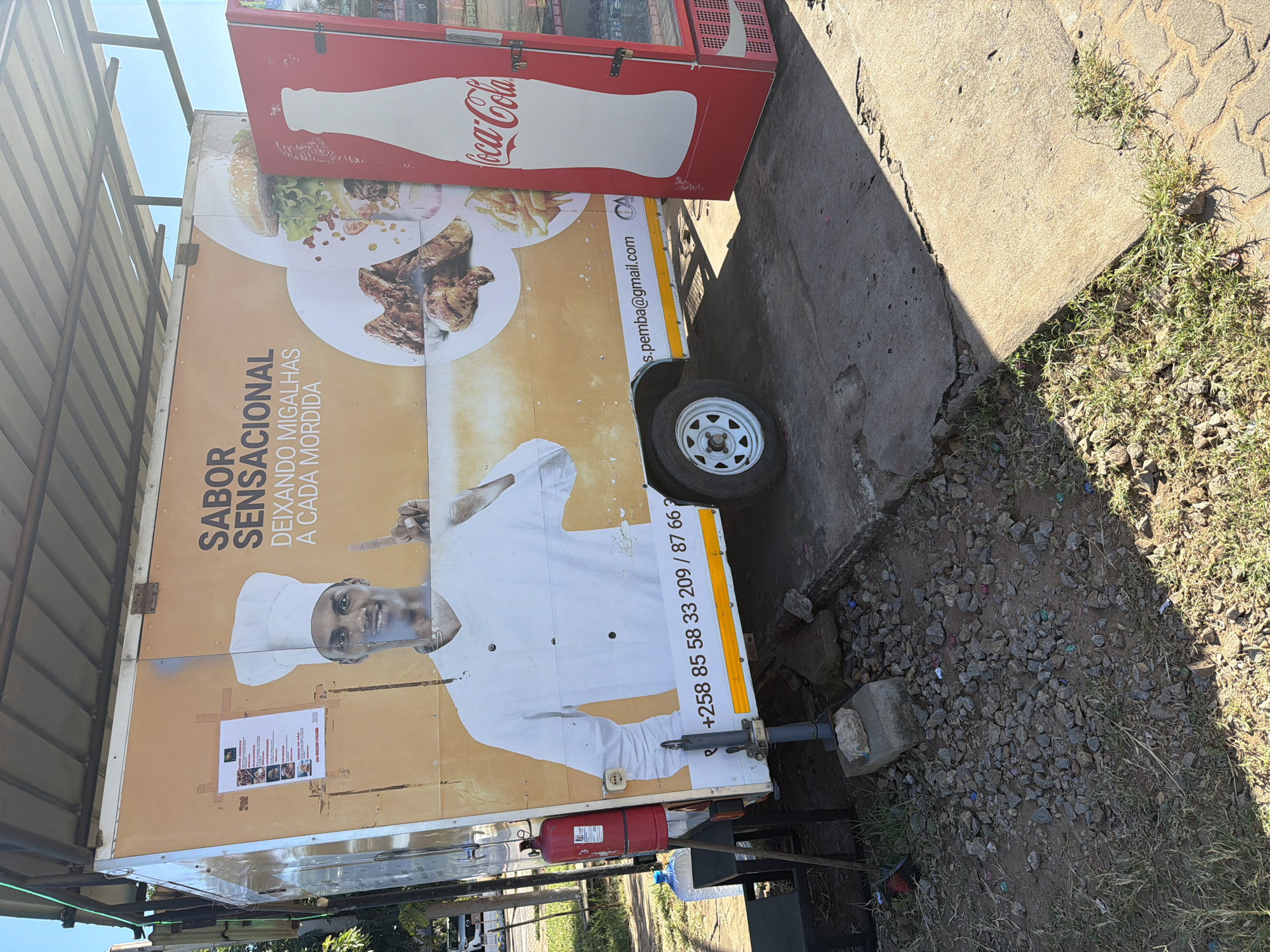 The image captures a mobile food truck or trailer parked outdoors under bright sunlight, with the scene rotated 90 degrees counter-clockwise. The truck is adorned with large graphics featuring images of food, including a burger, french fries, and grilled meat, along with a smiling chef wearing a toque, pointing to the text. A red Coca-Cola refrigerator, bearing the brand's iconic logo and bottle silhouette, is positioned adjacent to the truck.
The ground around the trailer is a mix of loose gravel, dirt, and patches of grass, with concrete slabs or curbs visible. Strong shadows cast by the objects indicate a sunny day. A small white wheel of the trailer is visible, as is a red fire extinguisher mounted near the hitch. A clear plastic water bottle also rests on the ground next to the truck. A small, printed document, likely a menu, is taped to the side of the truck, though its details are not clearly legible.
Visible text on the food truck includes: "SABOR SENSACIONAL" (Sensational Flavor) and "DEIXANDO MIGALHAS A CADA MORDIDA" (Leaving crumbs with every bite). Contact information provided is an email address: "s.pemba@gmail.com", and a partially visible phone number: "+258 85 58 33 209 / 87 66 7". The Coca-Cola logo is prominently displayed on the refrigerator. The overall scene suggests an outdoor food vending operation.