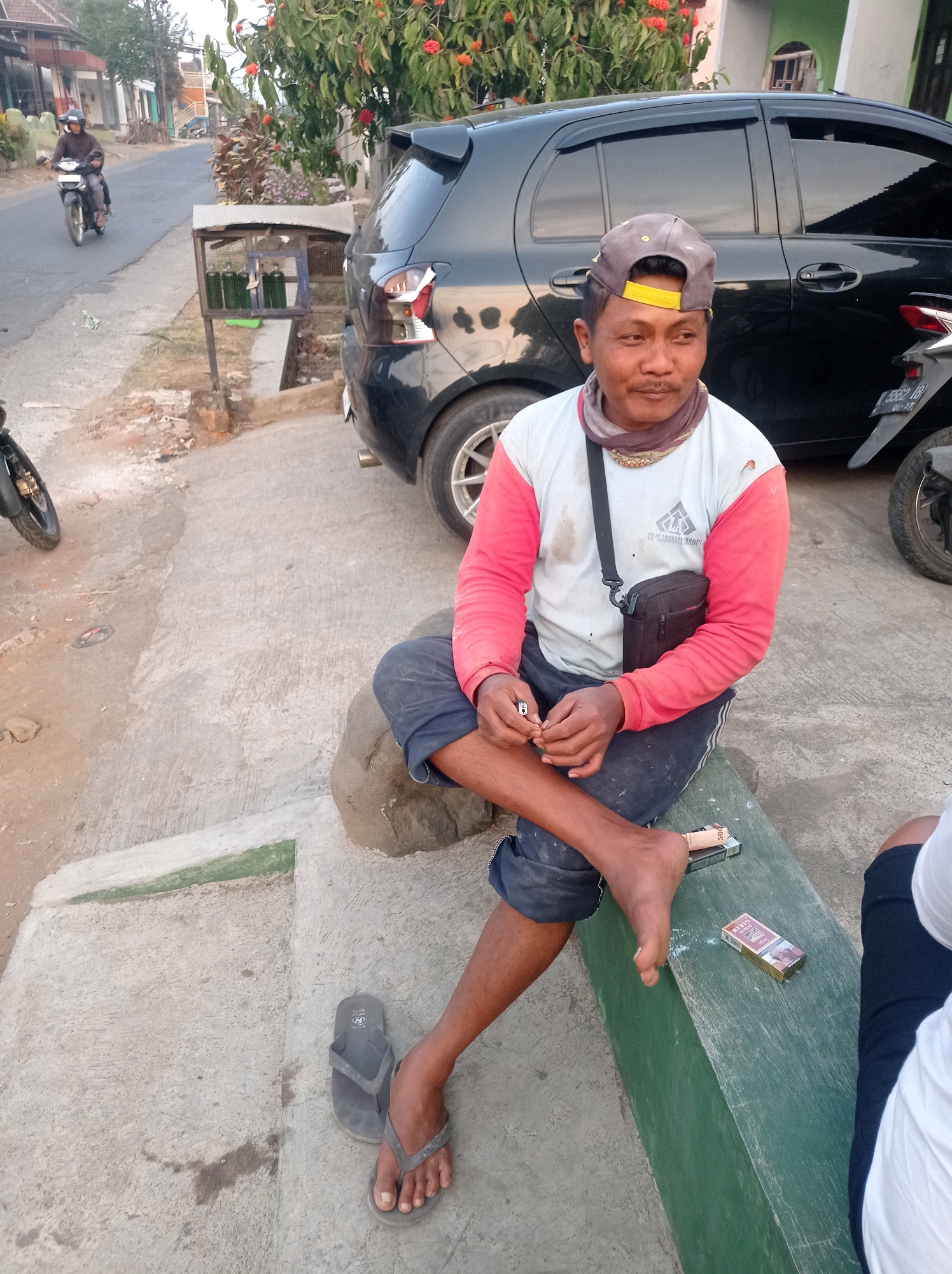 A man with a cap sits on a green bench in the city of Purwosekar, Indonesia, holding a cigarette. He wears a red-sleeved shirt and dark pants, with a small bag slung across his chest. Two packs of cigarettes rest on the bench near his feet. A black car is parked behind him, and a motorbike is visible in the background, along with a small structure holding what appear to be green bottles. The scene is set on a concrete path with a dirt shoulder, and vegetation with red flowers is present.
