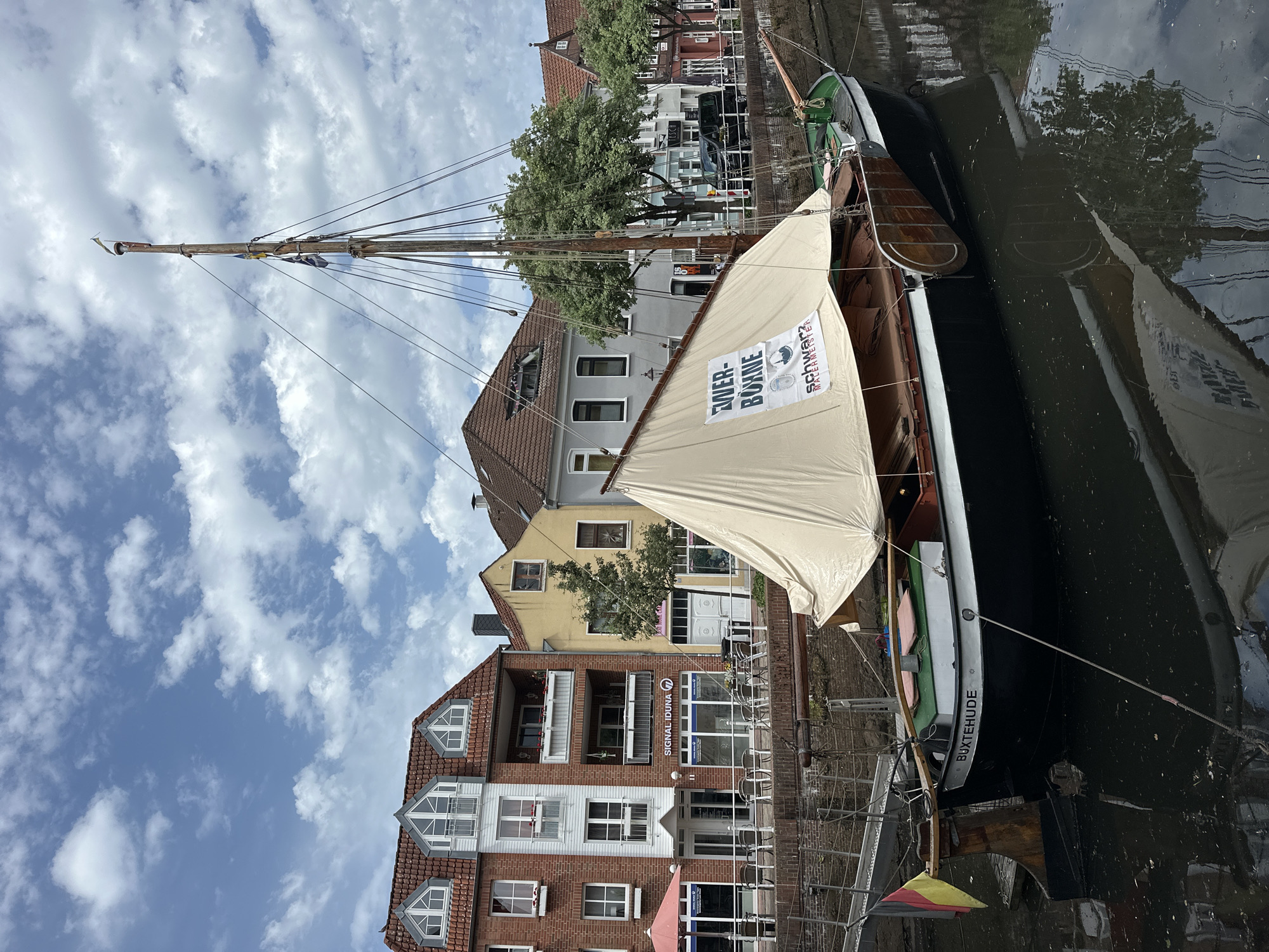 The image is taken in Buxtehude, Germany. A sailboat is docked on a canal, adjacent to a brick building with white trim and a storefront named "SIGNAL IDUNA". The sail is unfurled and bears the text "EUER BÜHNE" and "SCHWARZ MALERMEISTER". The boat's hull is black with "BUXTEHUDE" printed on it. Buildings with tile roofs and trees are visible in the background. The sky is partially cloudy.