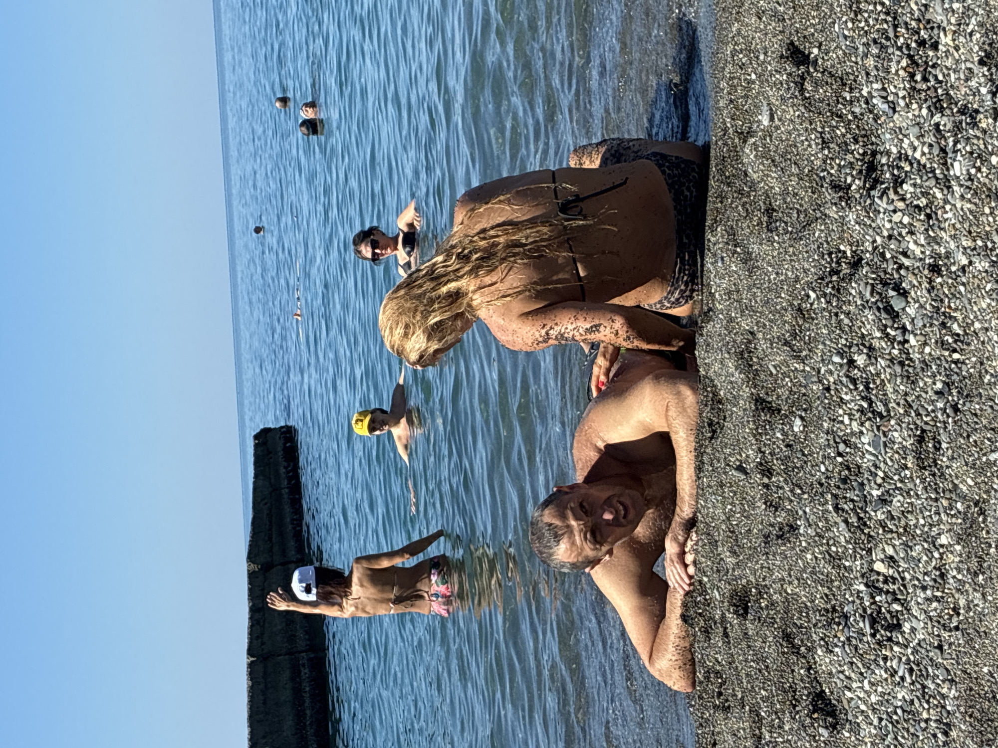 The image shows a sunny day at a beach in Sochi, Russia. In the foreground, a man is lying on the pebbly shore with his arms resting on the ground, looking at the camera. A woman with blonde hair, wearing a leopard-print bikini, is sitting on the shore behind him, facing away from the camera. Her back and arms appear to be covered in dark sand or mud. In the background, several people are in the blue water. One woman wearing a white cap is standing in the water with her arm raised. Another person wearing a yellow cap is also in the water. Further out in the sea, more people are visible. A concrete breakwater extends into the water on the left side of the image. The sky is clear and blue.