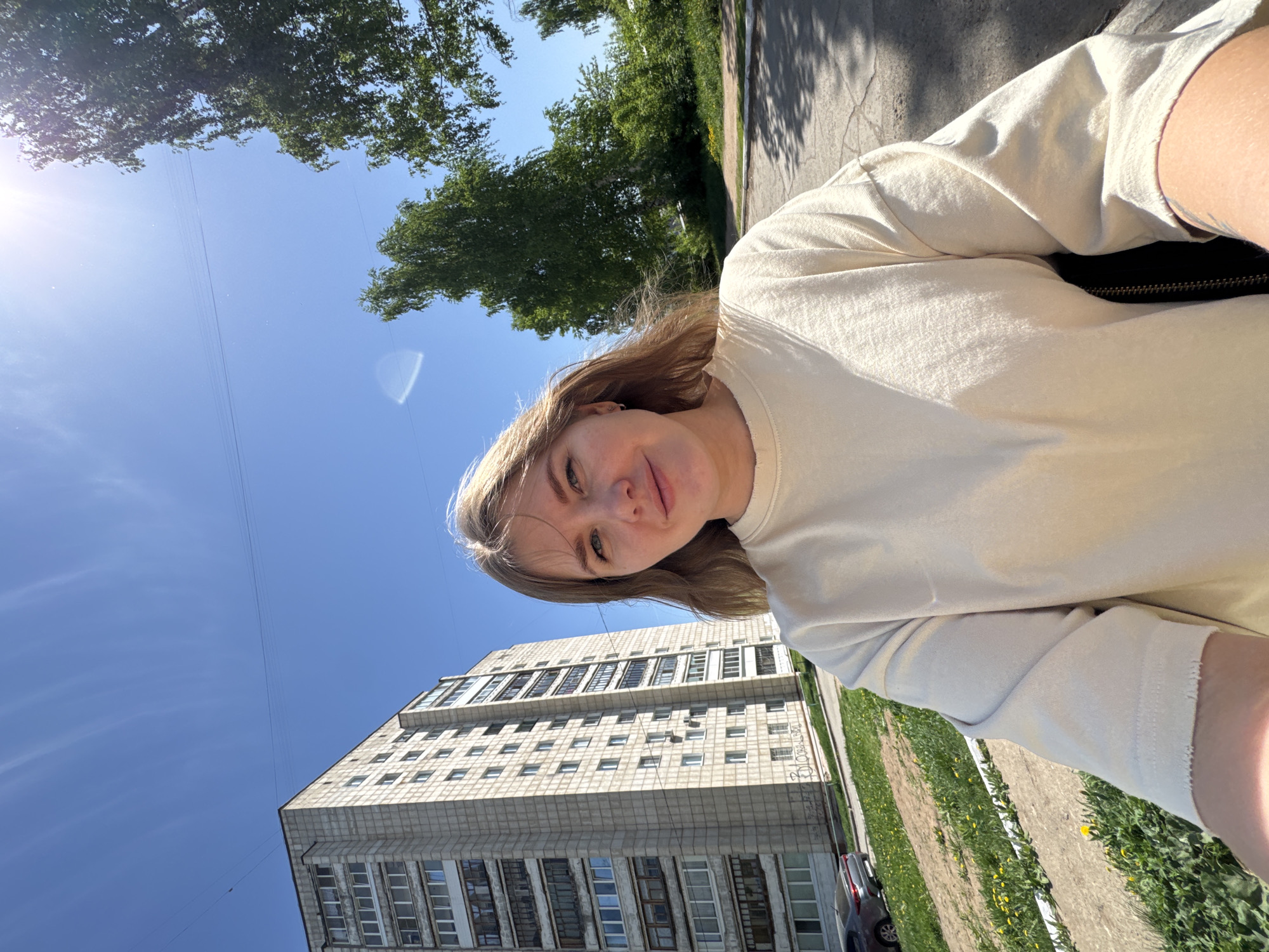 The image shows a selfie taken outdoors, likely in the city of Chaikovsky, Russia. The person is fair-skinned, with light brown hair and light-colored eyes. They are wearing a light-colored, long-sleeved shirt. Behind them is a tall, concrete apartment building with multiple rows of windows. The sky is blue with some trees visible at the top of the frame. There is a patch of green grass and a sidewalk near the apartment building. A vehicle is partially visible at the bottom right of the frame. The lighting indicates a sunny day.