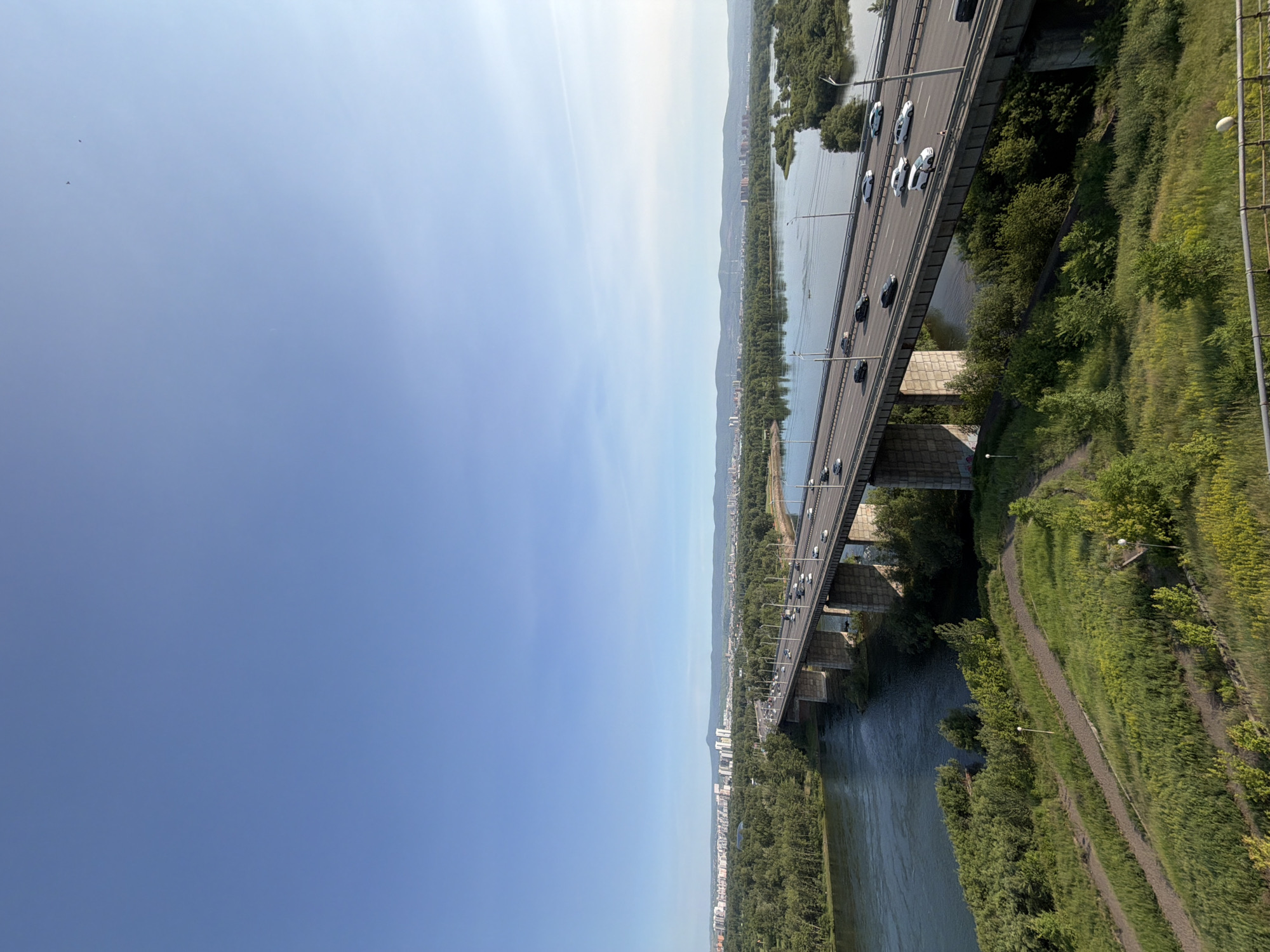 The media file shows an aerial view of a bridge spanning a wide river, with a city and mountains in the background. The image is oriented vertically, so the bridge and river run from the top left to the bottom right. The sky takes up the left half of the image and is a clear, bright blue, suggesting it is daytime and the weather is fair. A few small dark specks, possibly birds, are visible in the sky.
The bridge itself is a long, concrete structure supported by massive pillars that extend into the water. Cars, appearing as small dark and white shapes, are driving on the bridge, indicating that it is an active roadway. The river below is dark and calm, reflecting the sky and the surrounding greenery.
On the right side of the image, the riverbank is densely covered in lush green trees and vegetation, with a dirt path winding along it. This path is edged by more green grass and some low-lying yellow flowers. The overall scene suggests a natural, park-like setting on the riverbanks contrasting with the man-made infrastructure of the bridge and the distant city.
In the background, across the river, a city skyline can be seen with buildings of varying heights nestled among more trees. Beyond the city, a range of mountains rises, partially obscured by haze or distance. The scale of the bridge and the surrounding landscape implies a significant urban area.
No text is visible in the media file. The overall mood is one of a serene, somewhat expansive view, combining urban elements with natural scenery on a clear day. The location context of Krasnoyarsk, Russia, suggests this is likely a bridge over the Yenisei River.