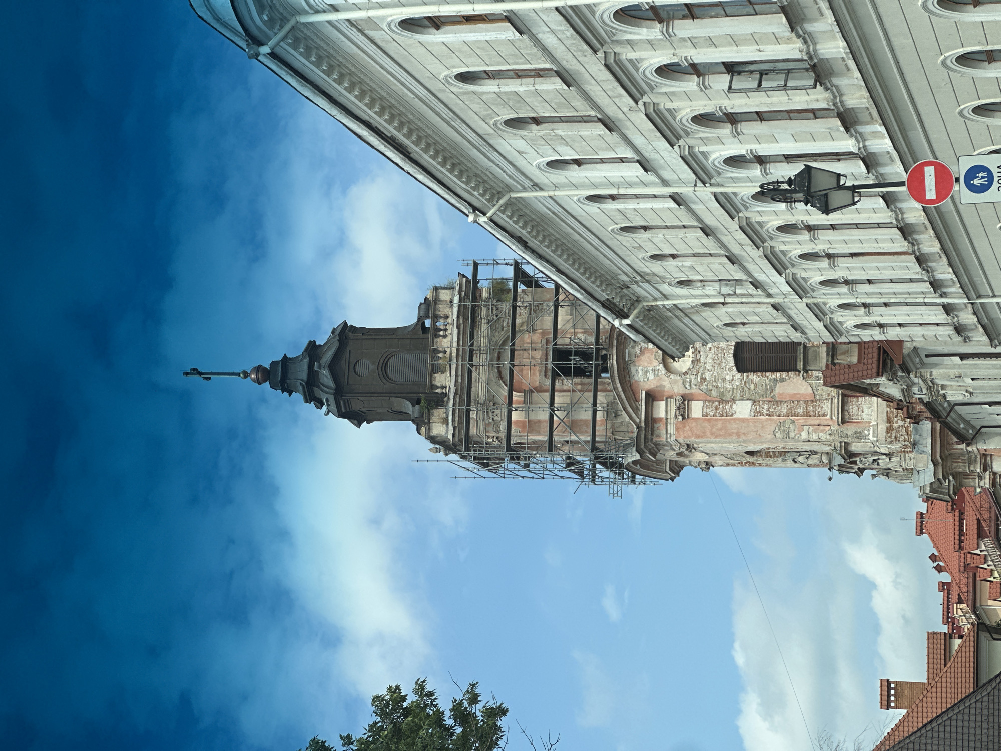The image shows a street view in Kamianets-Podilskyi, Ukraine, featuring a tall, old bell tower undergoing renovation, surrounded by scaffolding. To the right, a long, ornate building with numerous arched windows lines the street. The sky above is a deep blue with scattered white clouds. In the foreground, there are street signs, including a red and white prohibition sign and a sign depicting a pedestrian. The overall scene depicts an urban environment with historical architecture, suggesting a blend of the past and ongoing preservation efforts.