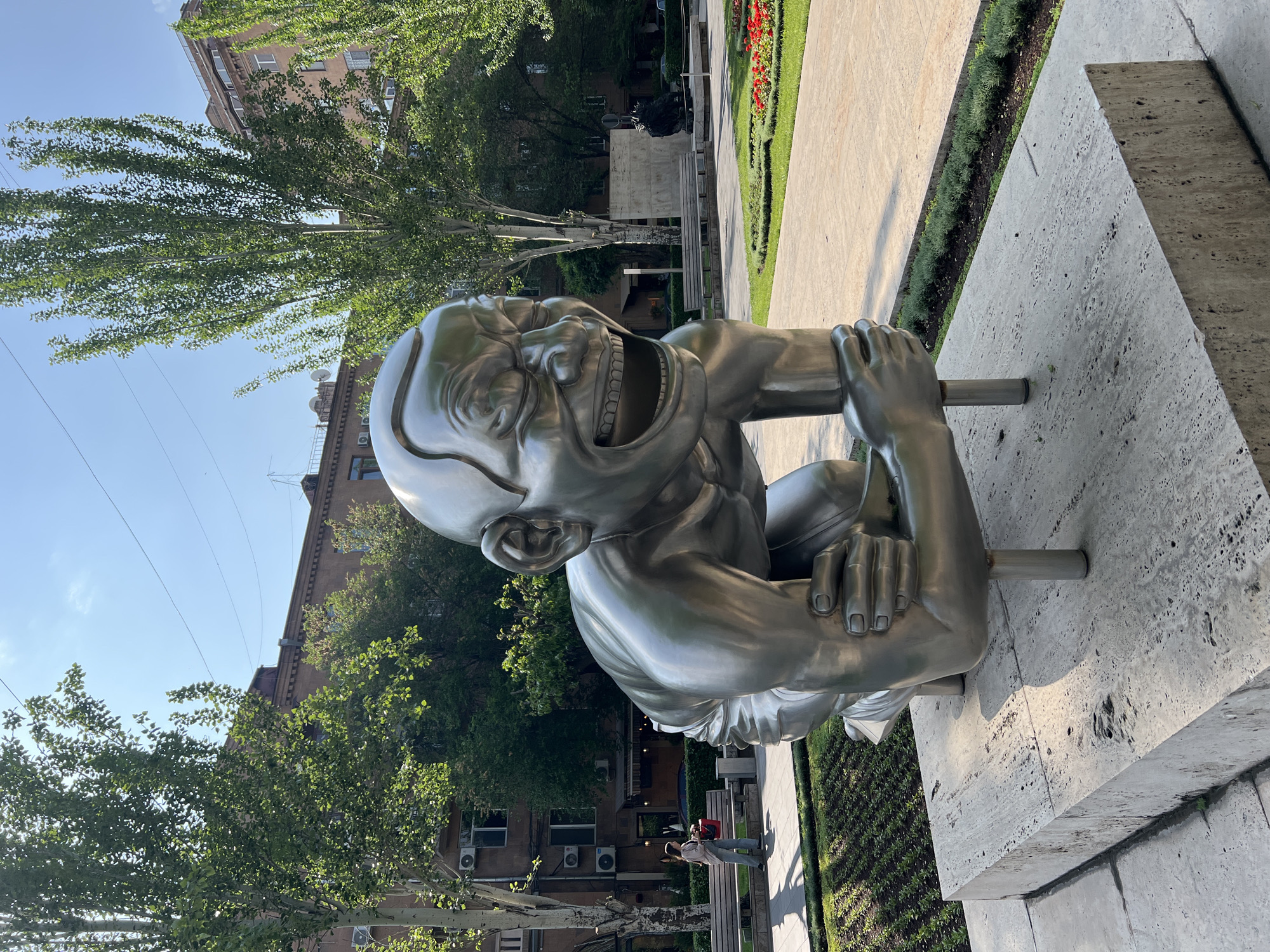 The image shows a large metal sculpture of a squatting figure with an exaggerated, open-mouthed grin. The sculpture appears to be part of a public art installation in Yerevan, Armenia, given the architectural style of the surrounding buildings and the landscaping. The figure is positioned on a concrete structure, and a park-like setting is visible in the background, complete with trees, flowers, pathways, and benches. In the distance, a woman and child walk alongside the walkway. The weather appears sunny.