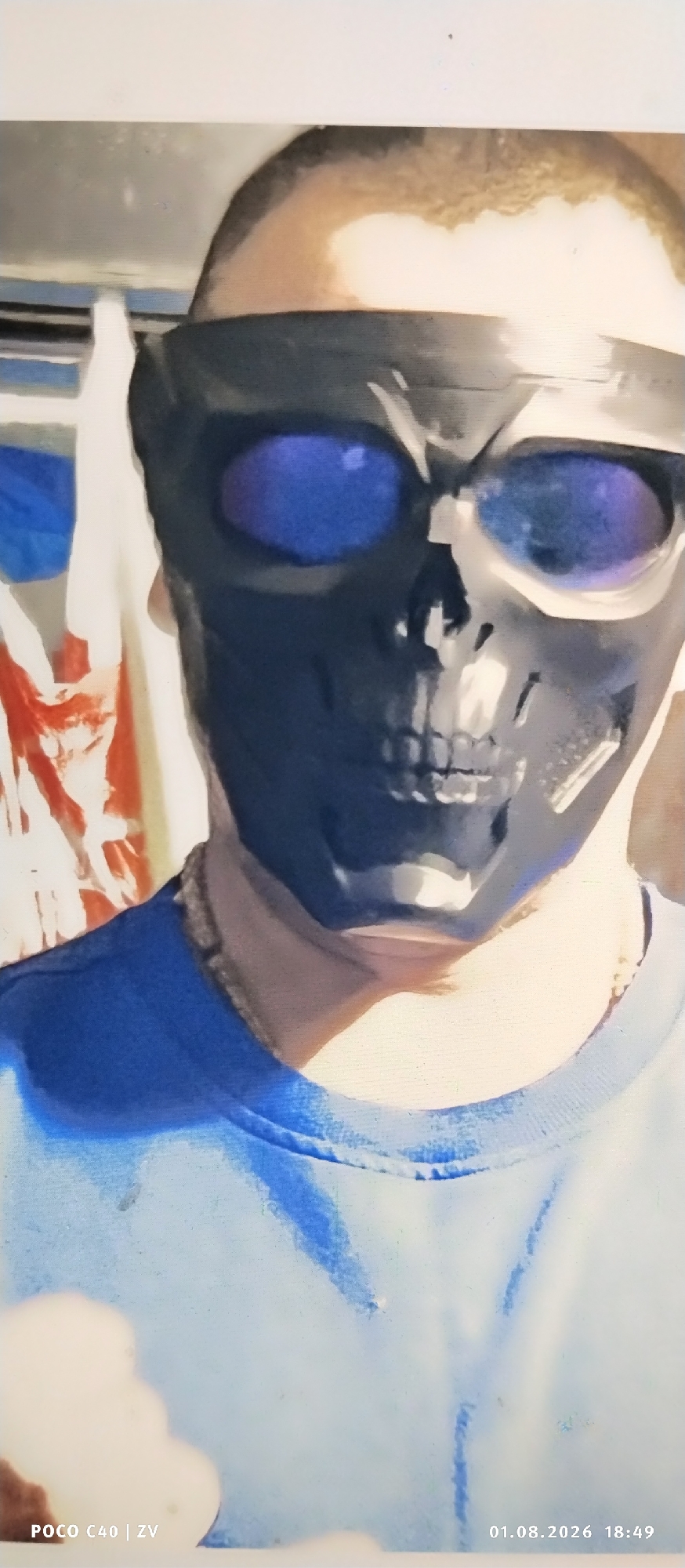 The image shows a close-up of a person wearing a black skull mask with blue lenses for eyes, and a blue t-shirt. The mask covers the entire face and has sculpted features resembling a skull, including teeth and eye sockets. The background is blurred, with hints of red and white patterns, possibly fabric or banners, and a lighter area that could be a wall or ceiling. The lighting is bright, casting some shadows on the person's neck and face. The bottom of the image includes text overlays: "POCO C40 | ZV" on the left and "01.08.2026 18:49" on the right, indicating the device and date/time the photo was taken. The image is cropped vertically, focusing on the upper body of the subject. There is no visible indication of the location being in Лубянки, Russia.