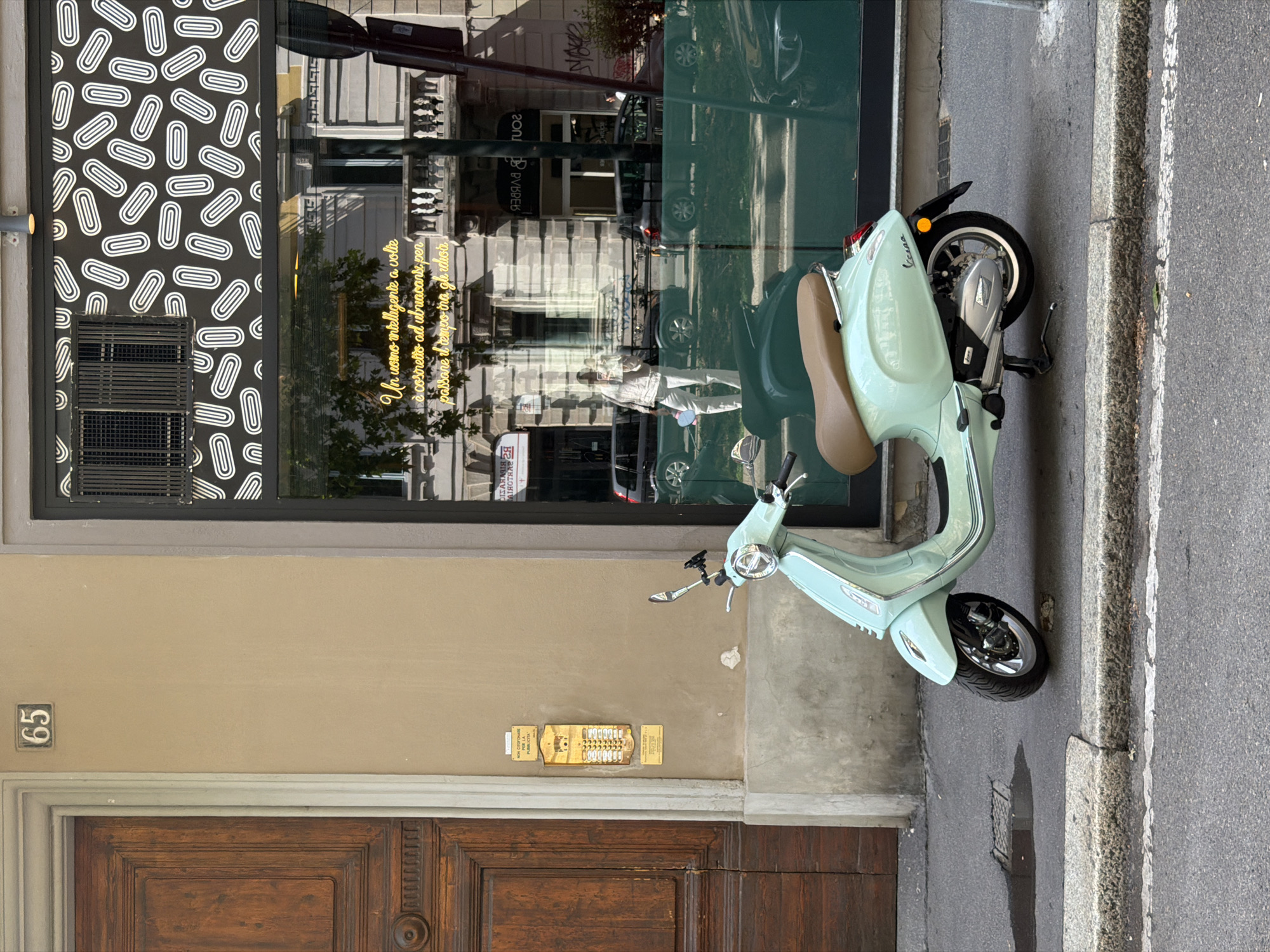 Here's a detailed description of the image:
**Overall Impression:** The image shows a mint-green Vespa scooter parked on a street in Milan, Italy. The scooter is situated next to a building with architectural and design elements typical of the city.
**Specific Details:**
* **Scooter:** The Vespa is the focal point. It's a classic Italian scooter, distinguished by its vintage design and light-green color. It appears to be well-maintained. The seat is brown.
* **Building Facade:** The building behind the scooter has a wooden door with "65" number on the wall. Above the door is an intercom system. A window above the door has a graphic pattern on the left and a reflection of the street across the road on the right.
* **Environment:** The street looks urban and has a typical sidewalk with curbing. There's a mix of light and shadow in the scene, suggesting either early morning or late afternoon light.
* **Location Clues:** The language of the text on the window, architectural style, and presence of the Vespa all strongly indicate Milan, Italy.
* **Quote on Window:** A yellow inscription on the window reads "Un uomo intelligente a volte è costretto ad ubriacarse per passare il tempo ina gli hor."