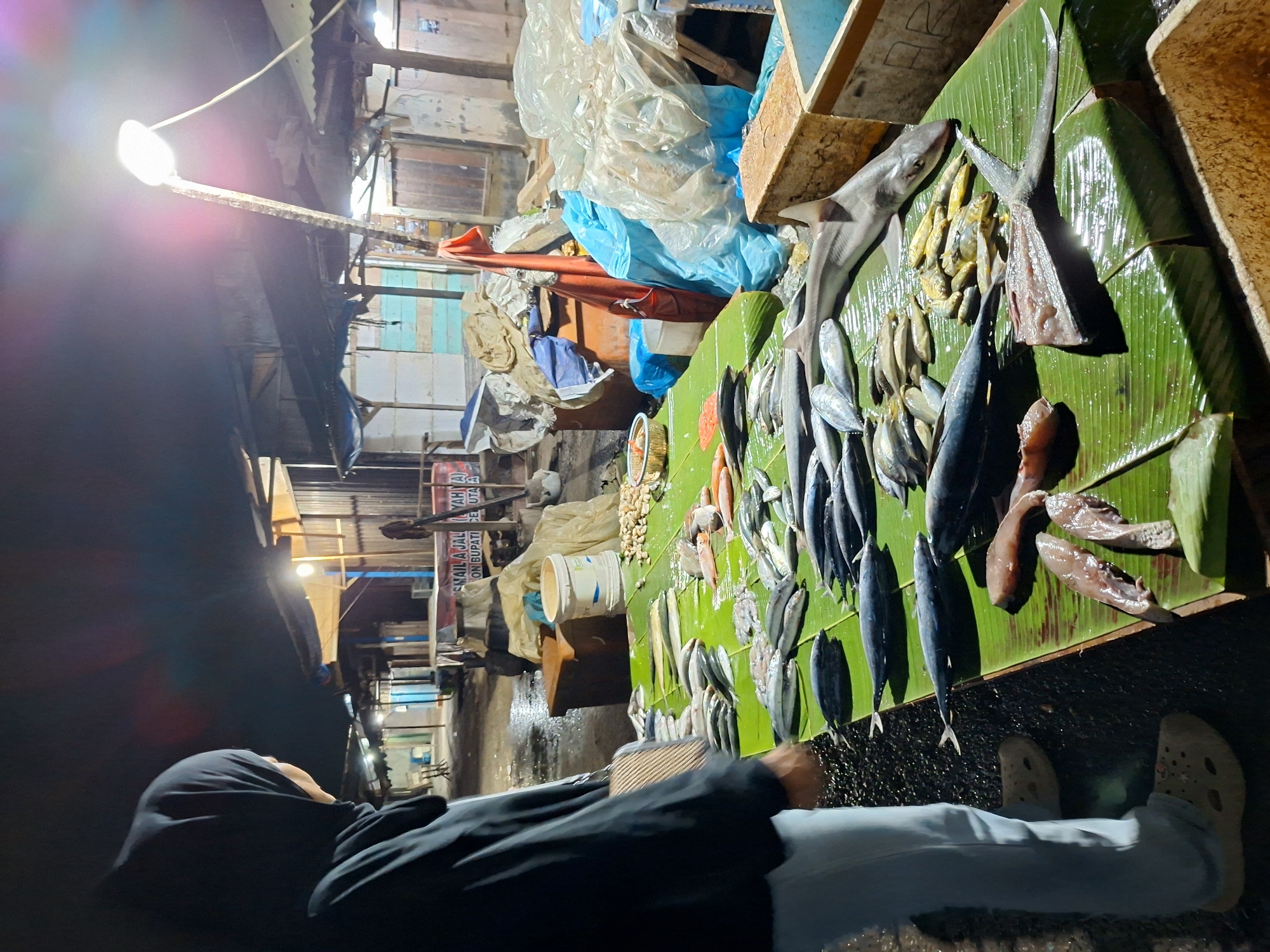 A street-level, medium shot depicts a woman standing in front of a fish market in Lhokseumawe, Indonesia. The woman is wearing a black hijab and a black long-sleeved shirt with light blue pants and beige Crocs. The market is illuminated by a bright overhead light, and displays a variety of fish laid out on green banana leaves. The fish include whole fish, fish fillets, and a shark. There are buckets and plastic sheeting near the fish display. Buildings and signs are visible in the background. The ground appears wet.