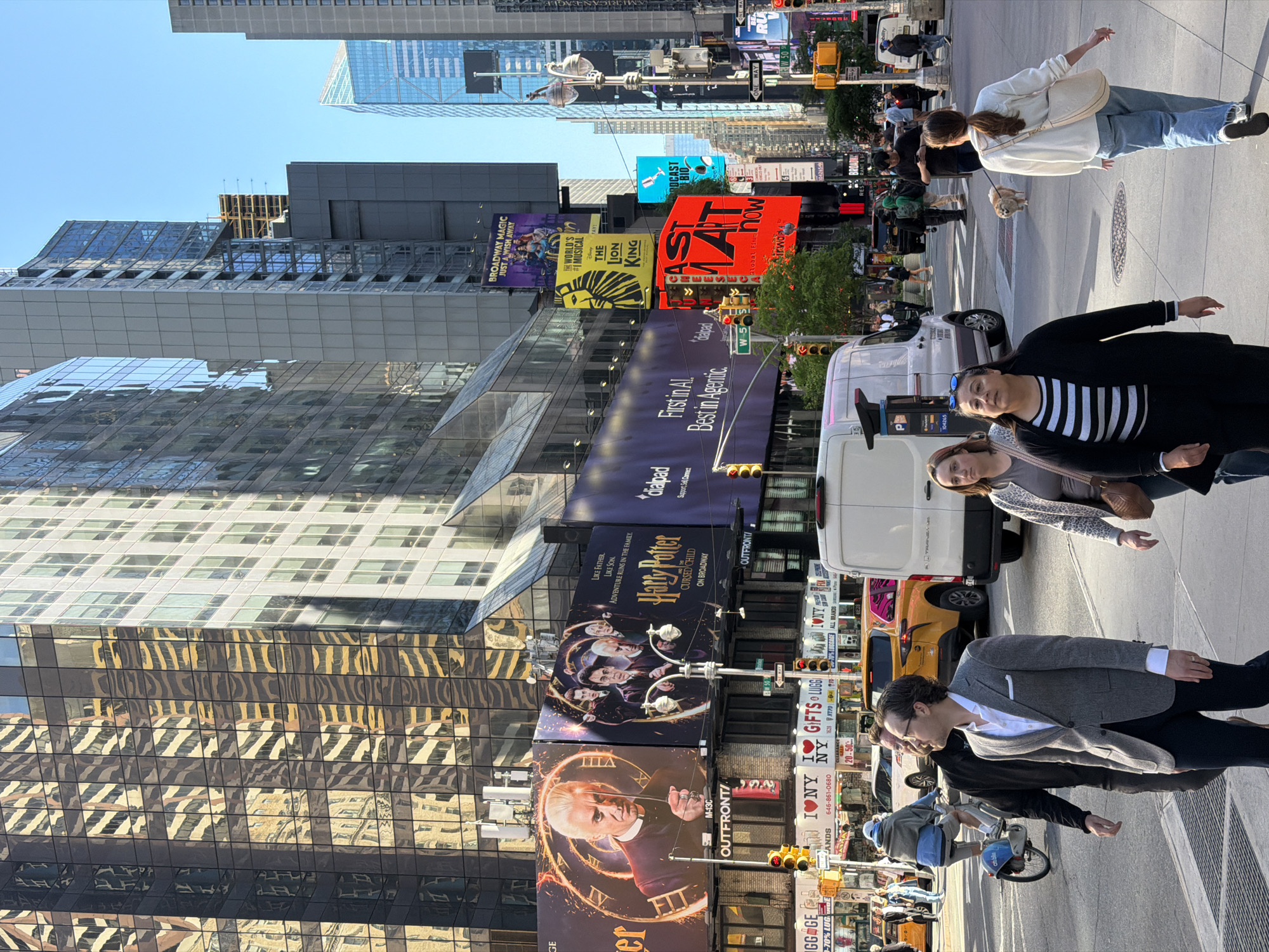 The image captures a vibrant street scene in Times Square, New York City, United States. Towering skyscrapers with reflective glass facades dominate the backdrop, adorned with large advertising billboards. Notable billboards include promotions for Broadway shows "Harry Potter and the Cursed Child," "The Lion King," and "Broadway Magic."
Pedestrians are present, including a woman with a dog, and other people on the crosswalks. Cars and vans can be seen interspersed among the pedestrian traffic. Street signs, traffic lights, and promotional advertisements are visible, including a luggage store.