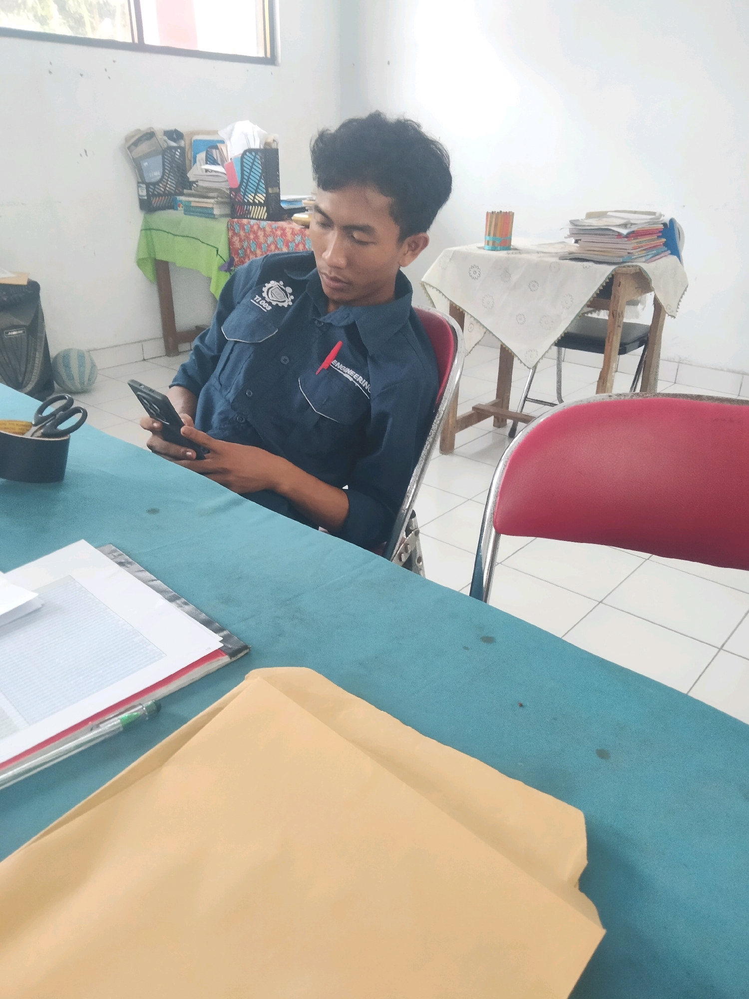 A young adult male, wearing a navy blue shirt with an embroidered emblem and the word "ENGINEERING" visible, is seated at a teal-colored table in what appears to be a classroom or study hall in Suka Sari, Indonesia. He is holding a black smartphone in his right hand, looking down at it. To his left, on the table, are a pair of black-handled scissors in a cylindrical holder, and a stack of papers or a binder. In the foreground, a tan manila envelope and a green and silver pen rest on the table. In the background, a wooden chair with a red upholstered seat is partially visible. Further back, a small wooden table is covered with a white tablecloth adorned with a circular pattern. On this table are stacks of books and a cylindrical container holding colored pencils. The room has white walls and a tiled floor. A window with a view of greenery is visible in the upper left portion of the image.