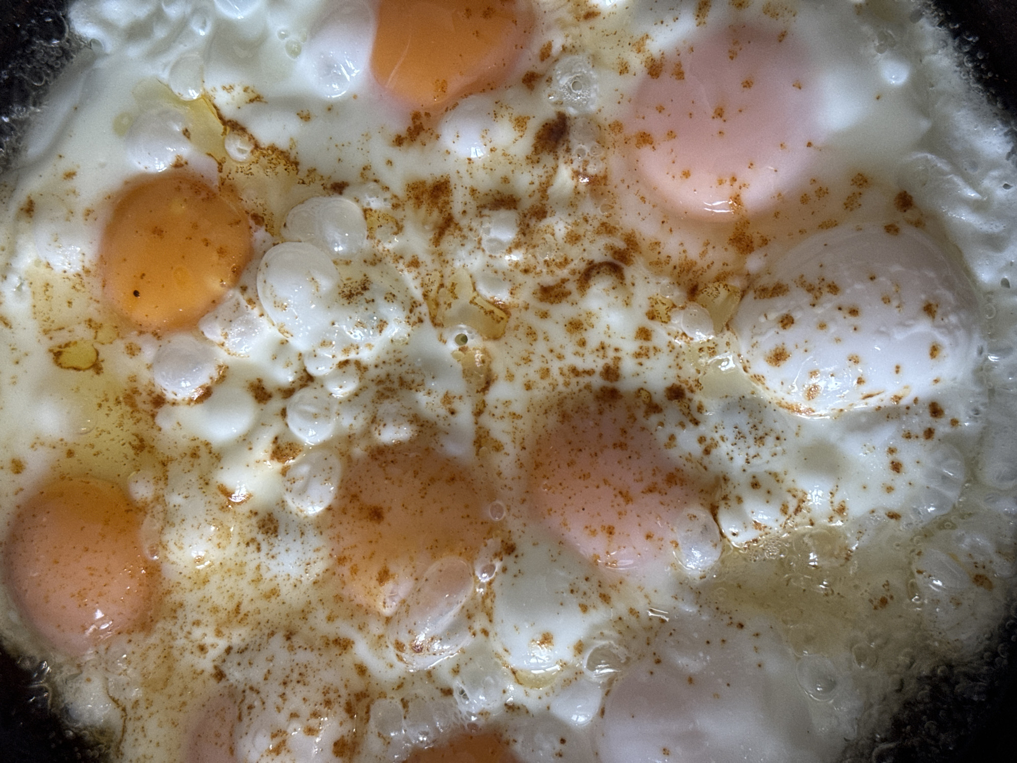The image depicts a close-up view of fried eggs in a pan. The eggs are mostly cooked, with varying degrees of firmness in the yolks, ranging from runny to slightly firm. The egg whites are mostly opaque and bubbly, with scattered browned bits. There is a concentration of browned seasoning or residue spread across the center of the pan. The edges of the pan are visible and appear dark, possibly from high heat or seasoning build-up. There is no information about this image being in Chaikovsky, Russia.