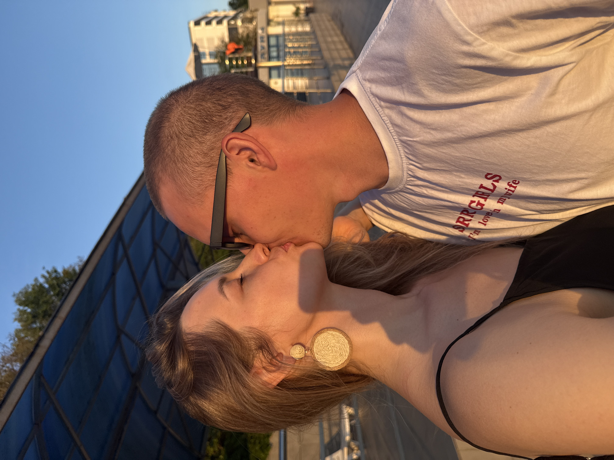 This image captures a close-up profile view of an adult couple, a man and a woman, kissing outdoors in Nebug, Russia, illuminated by warm, golden light from a low sun angle. The woman, on the left, has light brown hair and is wearing a black spaghetti-strap top. She has large, circular, woven straw earrings and her eyes are closed as she kisses the man. The man, on the right, has short-cropped hair, wears dark sunglasses on his head or eyes, and a white crew-neck t-shirt with red lettering on the chest that partially reads "SRRGIRLS" and "I'm love h my wife". Their lips are touching as they face each other. In the background to the left, a blue, translucent canopy or roof structure with metal framing is visible, with green tree foliage behind it. To the right, a clear blue sky is visible above a multi-story, light-colored building with multiple windows and balconies. A metal railing and an orange, possibly traffic-related, object are also discernible in the mid-ground on the right.